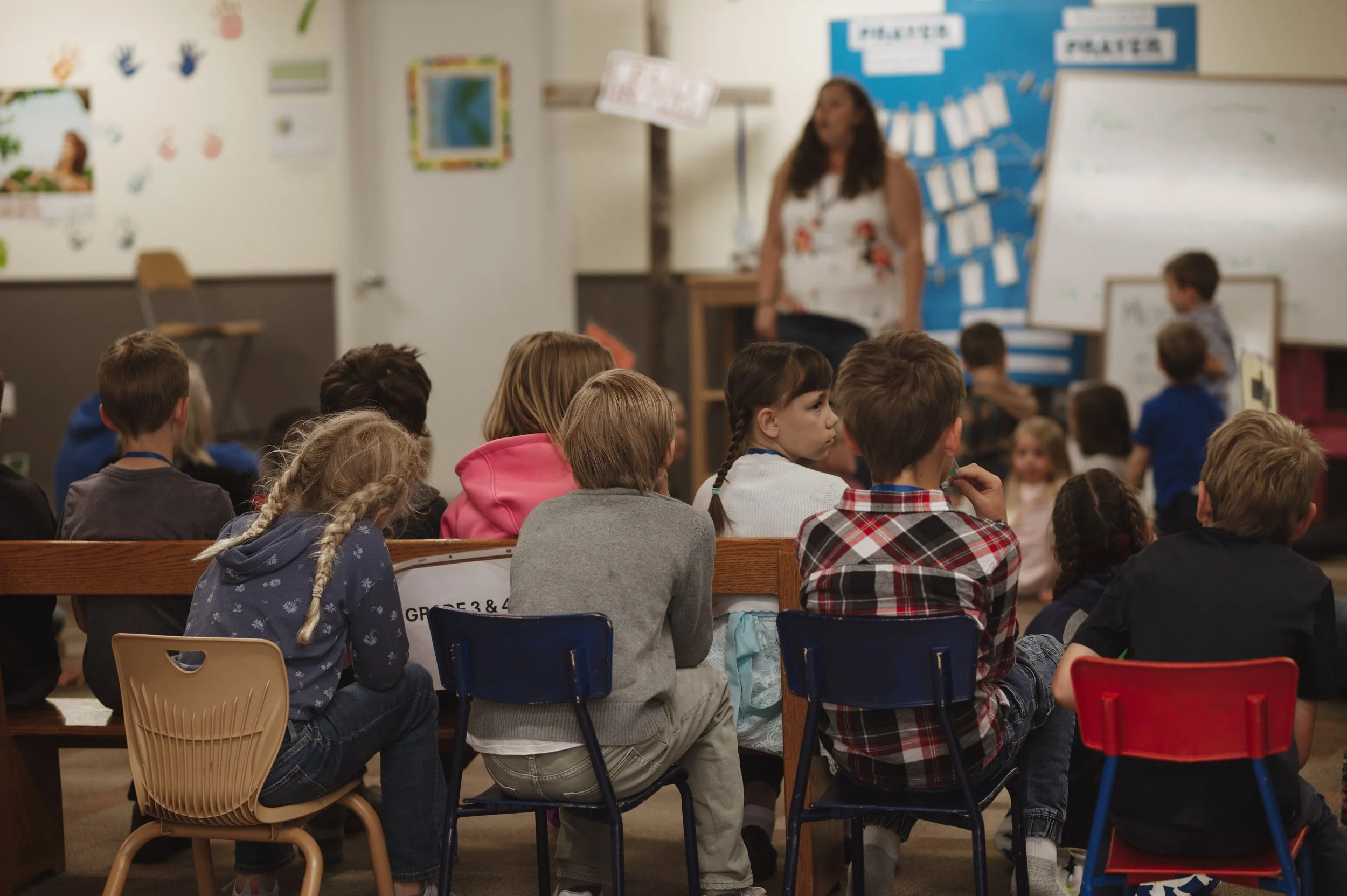 School classroom scene with children sitting on chairs facing a whiteboard; teacher in front; colorful classroom decorations on walls.