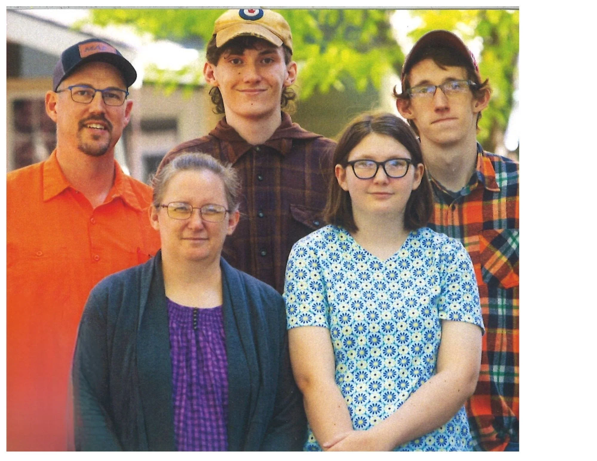 A group of five people standing outdoors, smiling, with trees and blurred background. The group includes three men and two women, with casual attire and glasses.