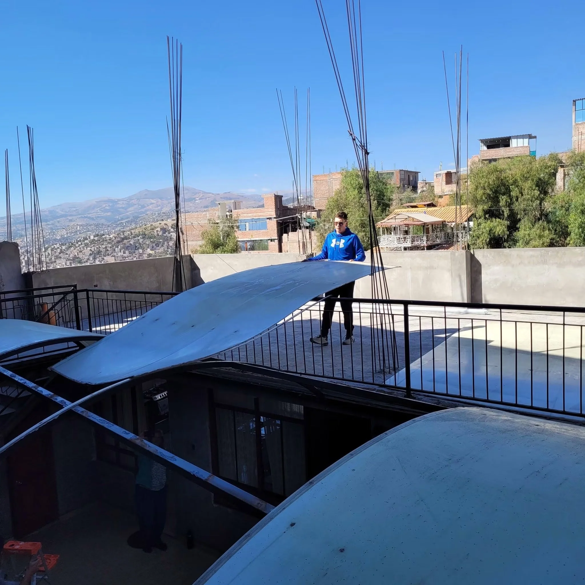 A man wearing sunglasses and a blue hoodie standing outdoors in front of a construction site with metal beams and buildings, with mountains and a clear blue sky in the background.