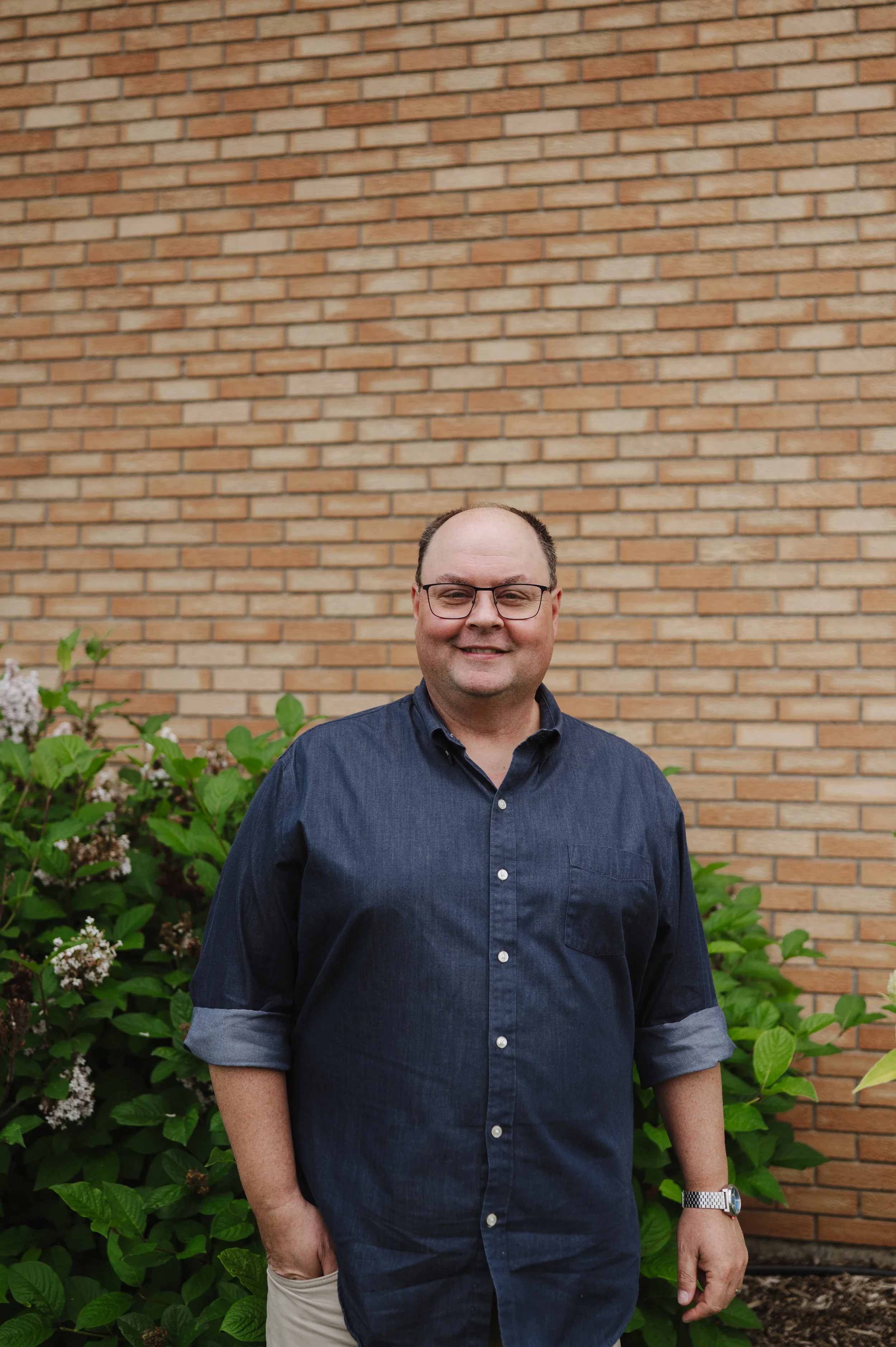 A man with glasses and a watch standing outdoors in front of a brick wall and green bushes, smiling at the camera.