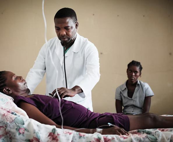 A male healthcare worker in a white coat checking a female patient's pulse with a stethoscope in a hospital room, with another woman sitting in the background.