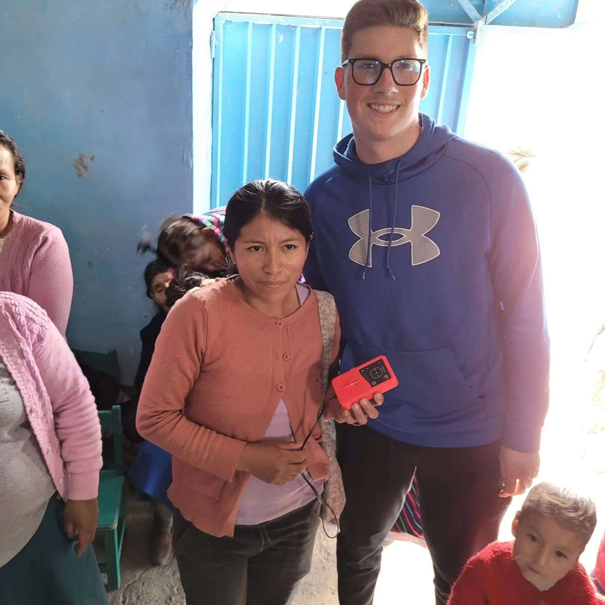 A group of people, including children and a young man wearing black glasses and a blue hoodie, some smiling, standing outside near a blue wall and blue door, with sunlight shining.