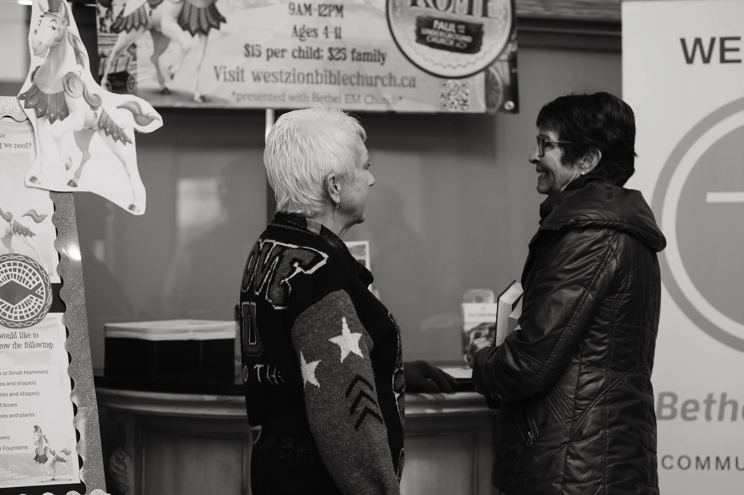 Two women face each other and are engaging in conversation while smiling. One woman has short white hair and is wearing a patterned sweater, and the other woman has short dark hair, glasses, and is wearing a jacket. The background contains posters and signages.