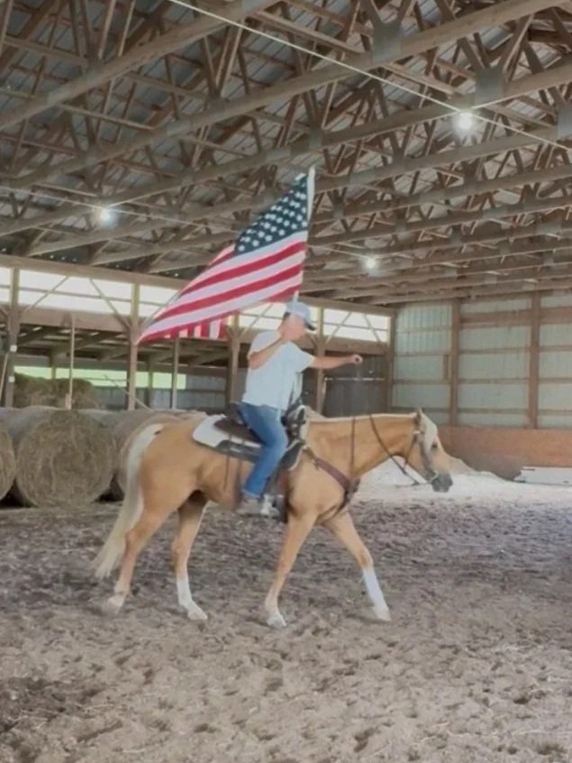 Person riding a horse indoors, holding an American flag attached to a pole, wearing a cowboy hat, white shirt, and jeans.