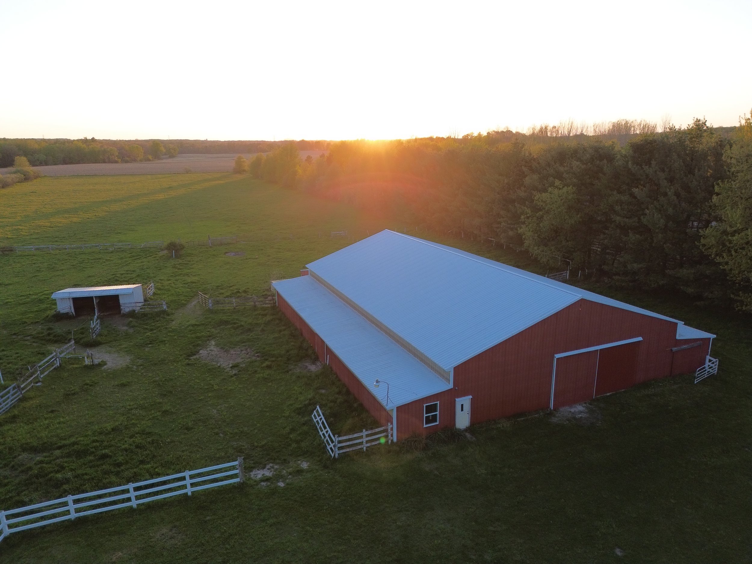 A barn with a silver roof and white fences on a green farm, with trees and fields in the background under a sunset sky.