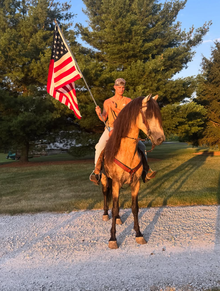 Young man riding a horse outdoors, holding an American flag, in an open grassy area surrounded by trees.