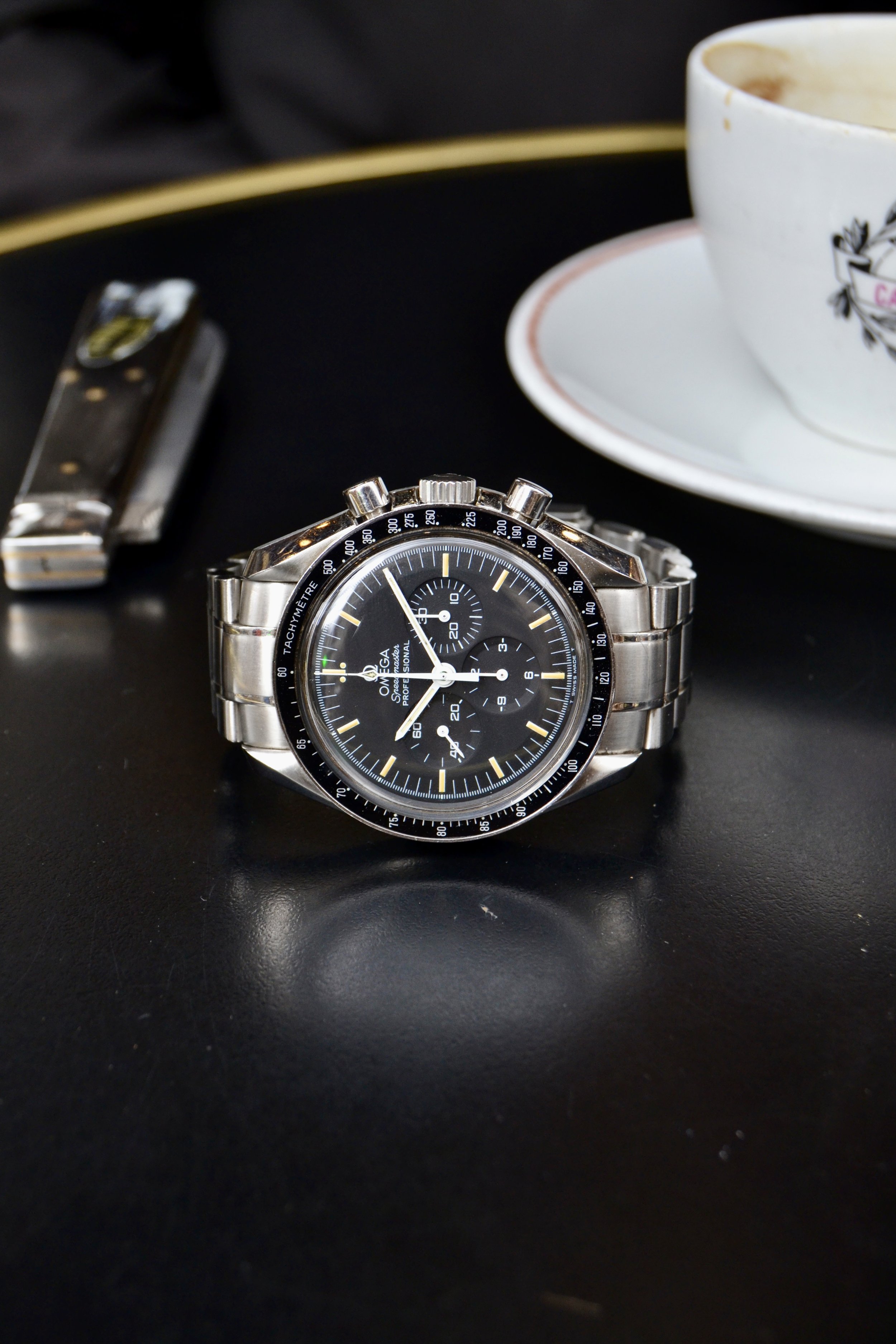 Omega Speedmaster en acier inoxydable avec cadran noir, chronographe et tachymètre sur un fond noir avec une tasse et une soucoupe blancs en arrière-plan.
