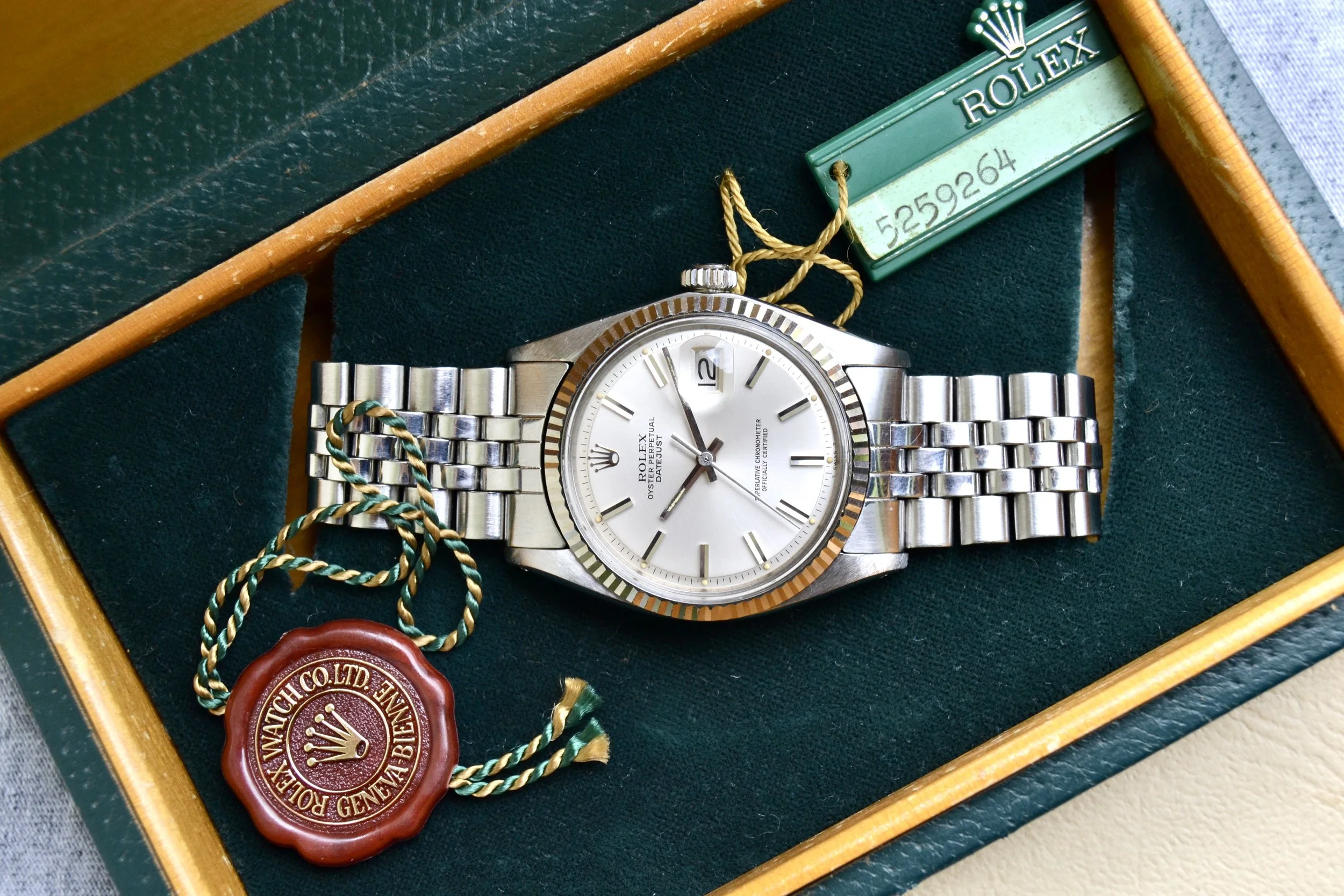 Montre Rolex en acier argenté avec cadran blanc, présentée dans une boîte en bois verte avec intérieur en velours vert foncé, accompagnée d'une étiquette de vente Rolex et d'un sceau en plastique rouge de la marque Rolex.
