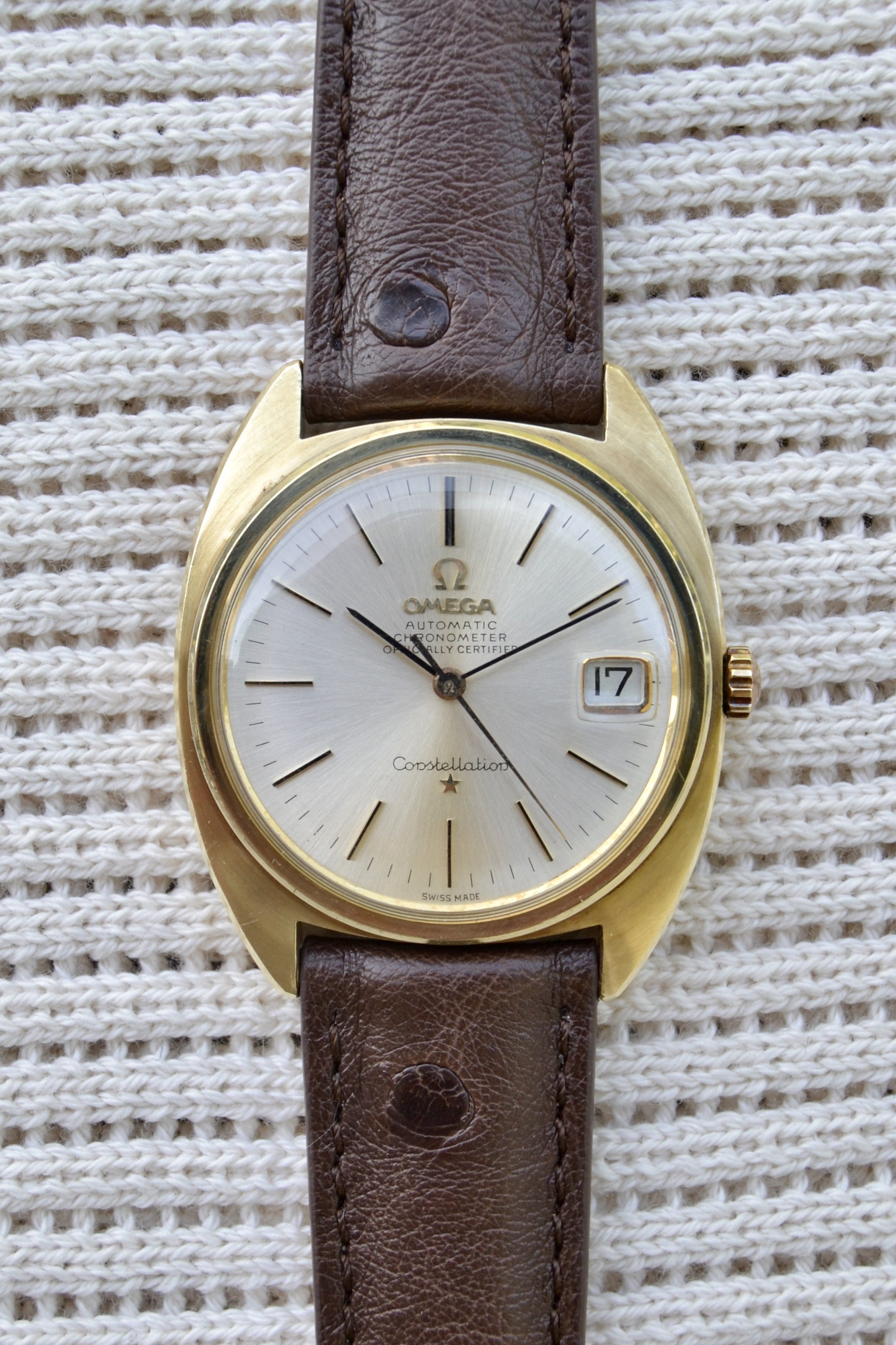 Montre Omega avec un bracelet en cuir marron, cadran argenté, affichant la date 17 et des aiguilles noires, posée sur un tissu tissé blanc.