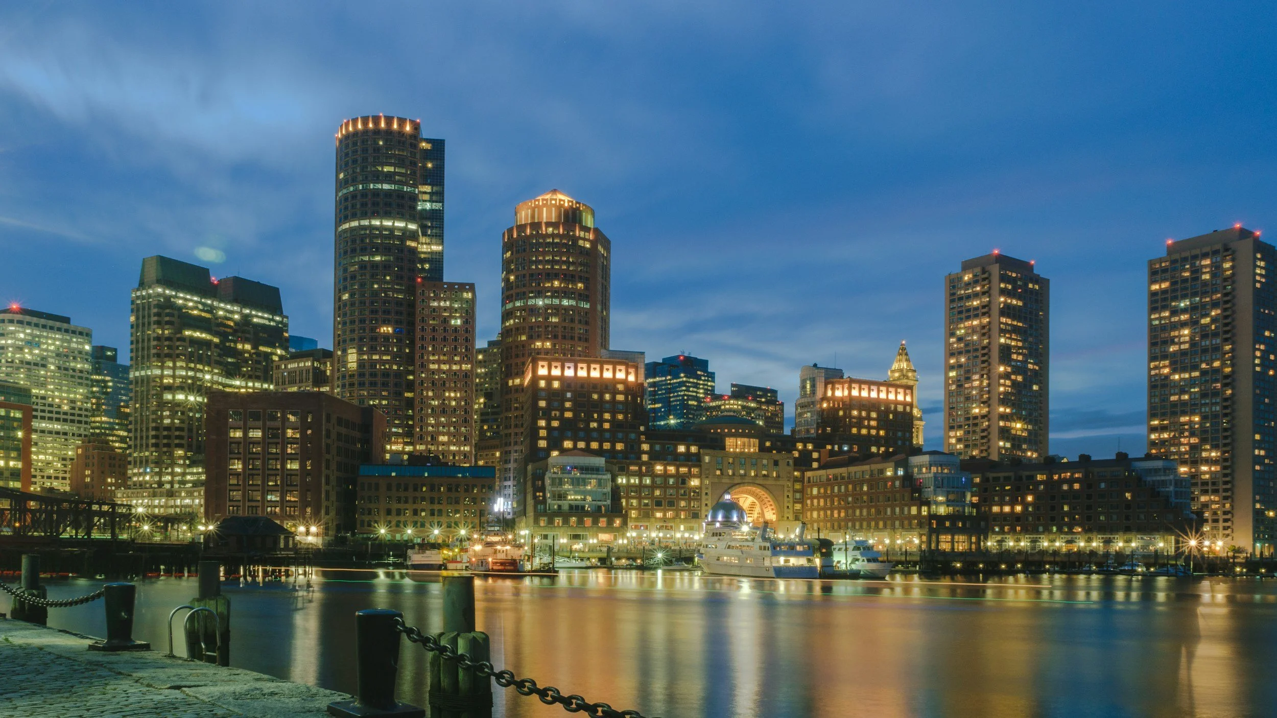 Nighttime view of a city skyline with tall illuminated buildings, a waterfront with boats, and calm water reflecting city lights.