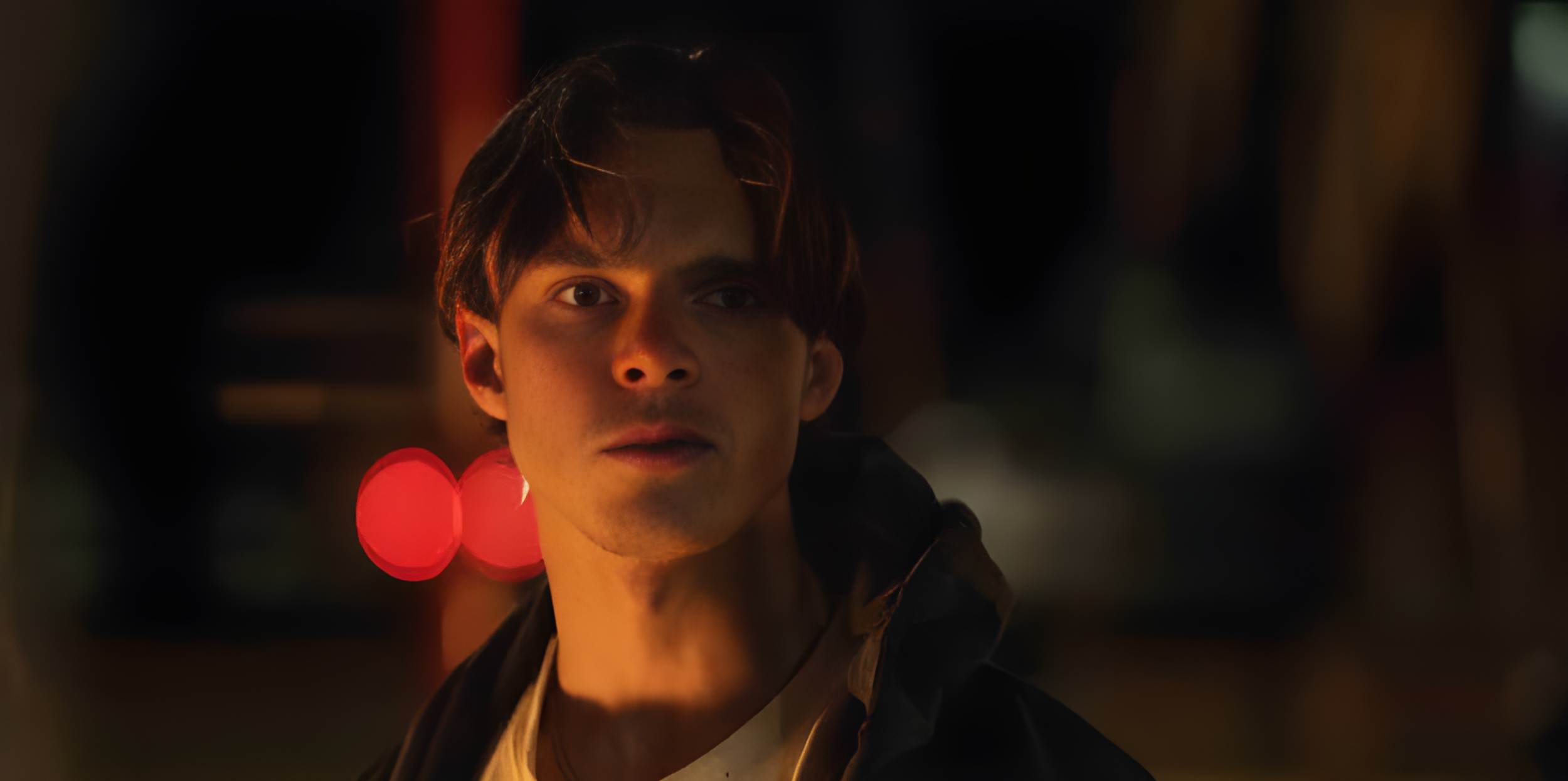A young man with dark hair and light skin is illuminated by a warm light, with blurred background lights in red and green. He is wearing a dark jacket and a beige shirt, looking intently to the side.