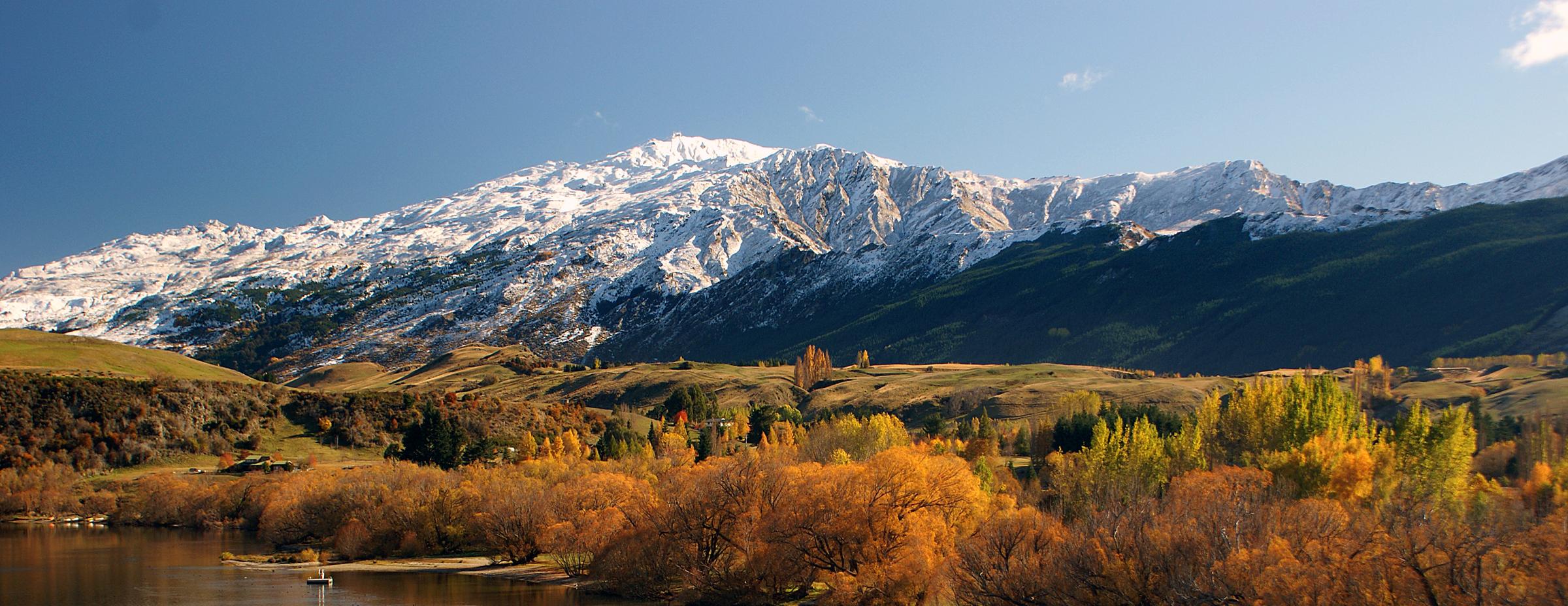 Snow-capped mountains beyond rolling green hills and colorful autumn trees near a body of water.