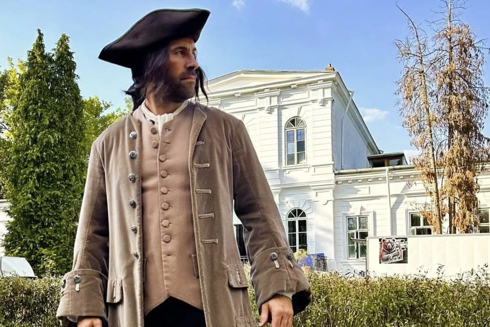 A man dressed in historical colonial attire, wearing a tricorn hat, stands outdoors near a white building with arched windows and greenery, under a blue sky with some clouds.