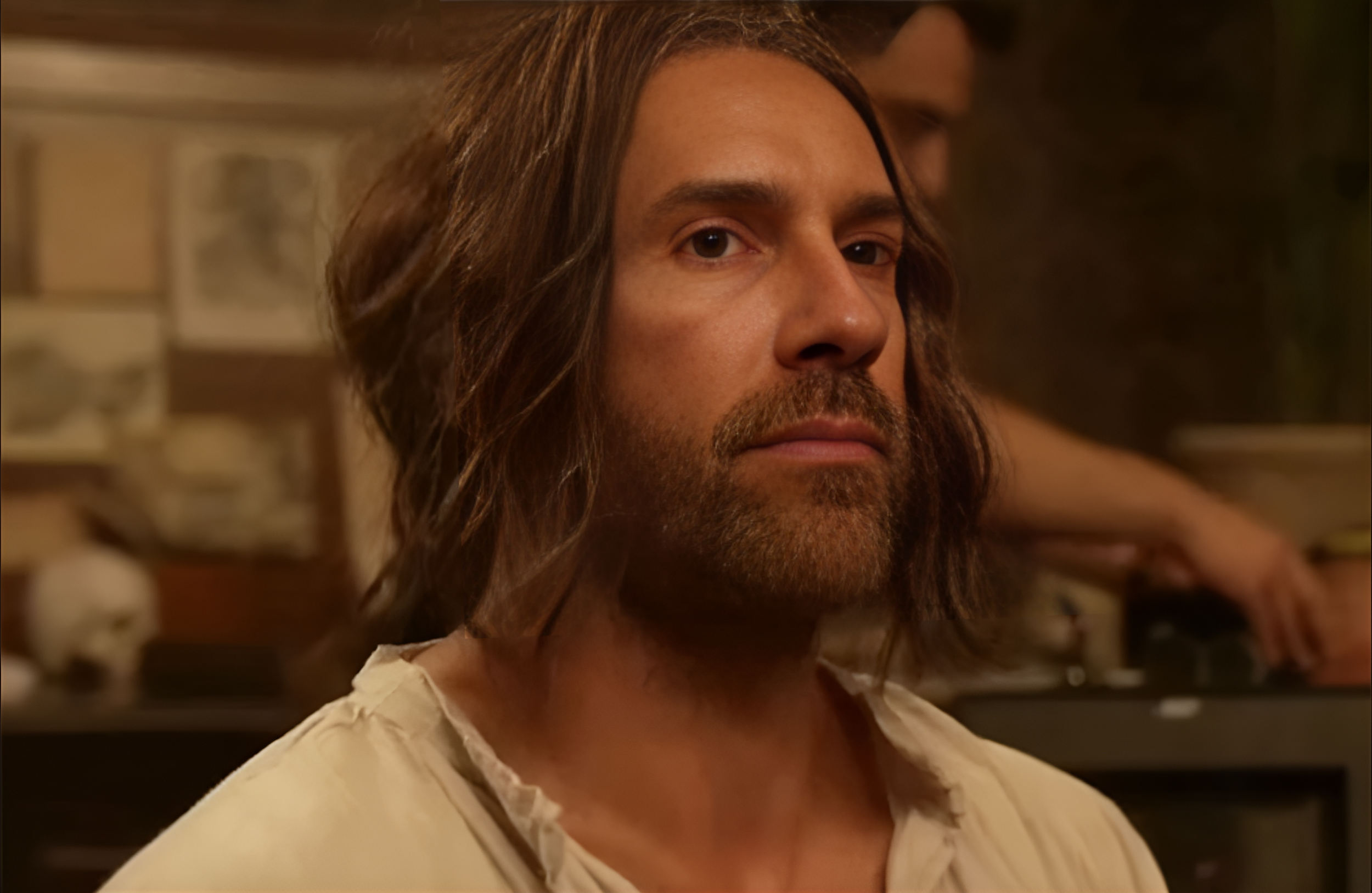 Close-up of a man with long hair and a beard, wearing a torn cream-colored shirt, standing in front of a brick wall with skulls on shelves.
