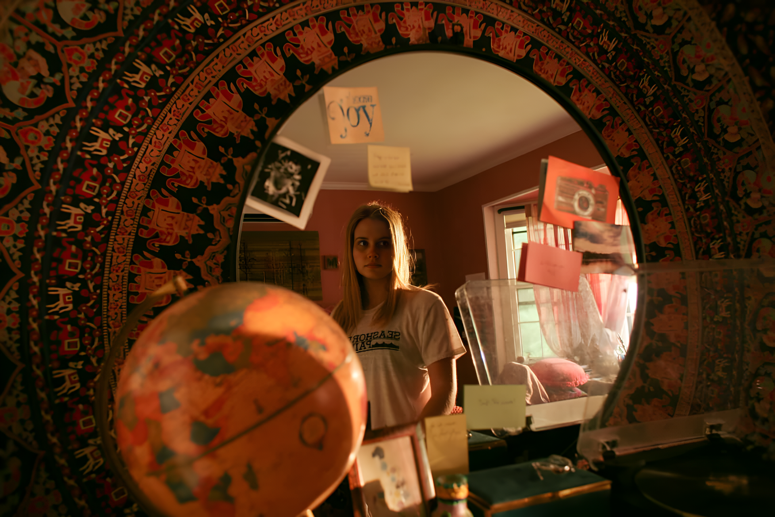 A young woman with long hair looking into a mirror with a colorful border in a room decorated with hanging notes, posters, and artwork. The room has warm lighting and a window letting in natural light.