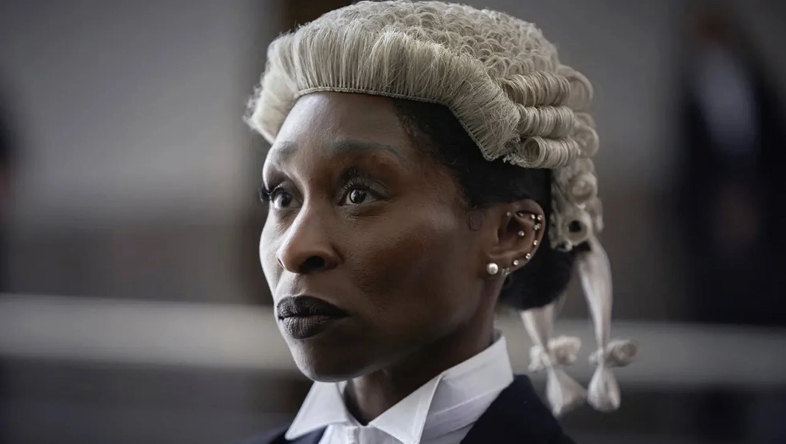 Close-up of a woman with styled gray hair, various piercings in her ear, dark lipstick, and wearing formal attire, with a neutral background.