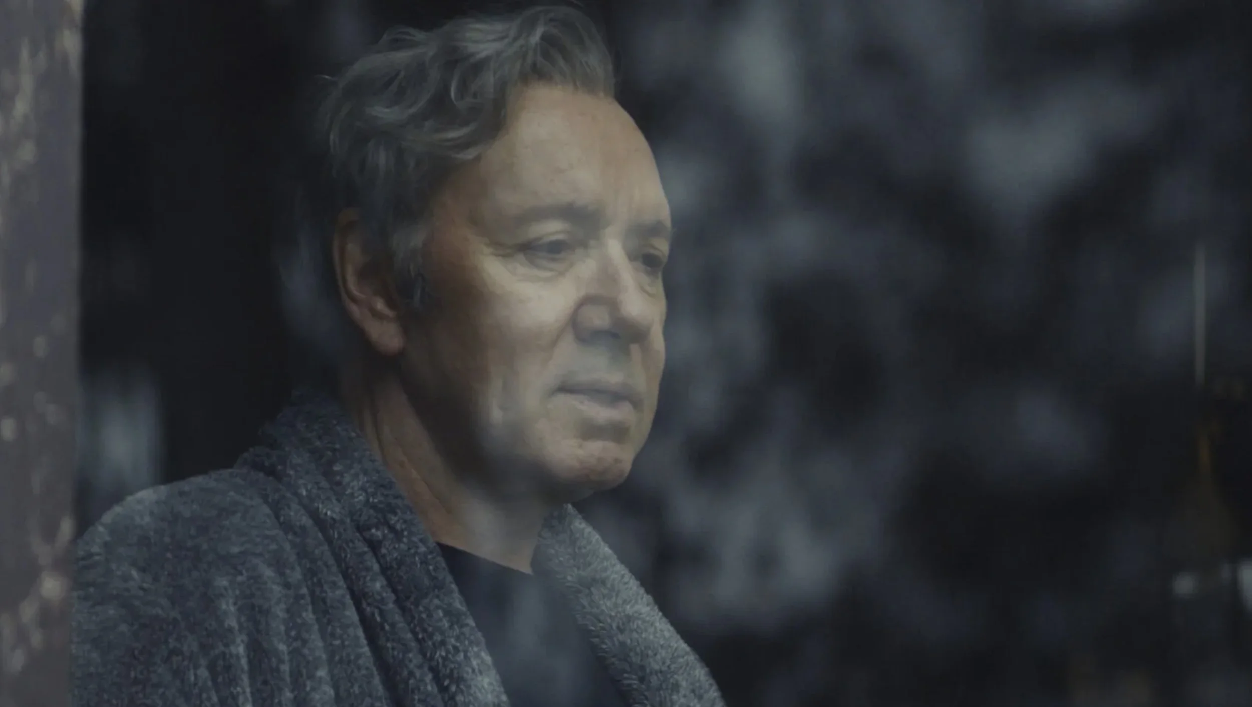 Close-up of an elderly woman with short gray hair, gazing thoughtfully outside through a foggy window.