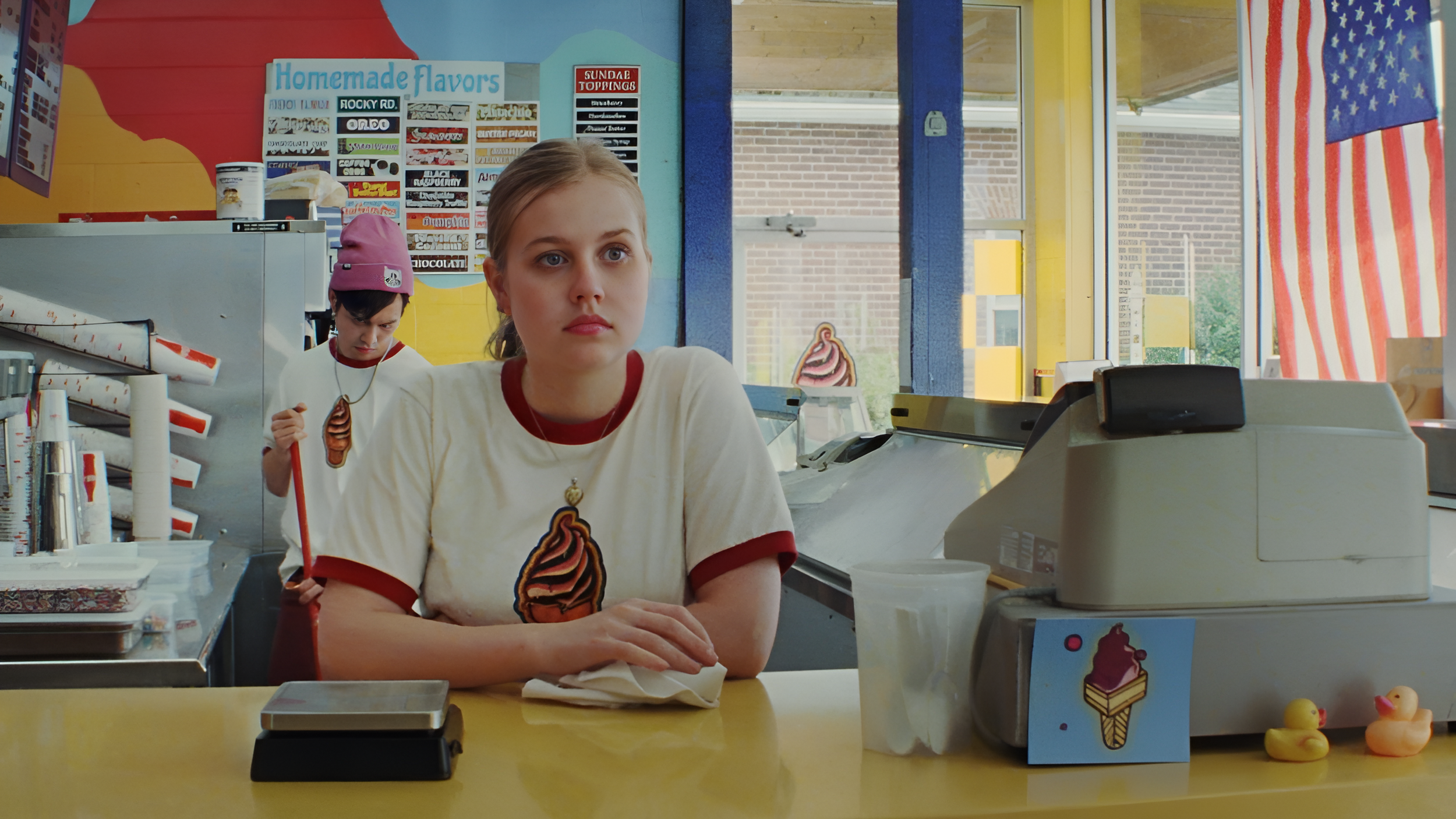 A young woman standing behind a counter at an ice cream shop, with a young man in the background. The woman is wearing a cream-colored T-shirt with an ice cream cone graphic, and the man is wearing a pink beanie. The shop has colorful walls and a "Homemade Flavors" sign with various toppings listed. There is an ice cream machine in front of the woman and rubber duckies on the counter.