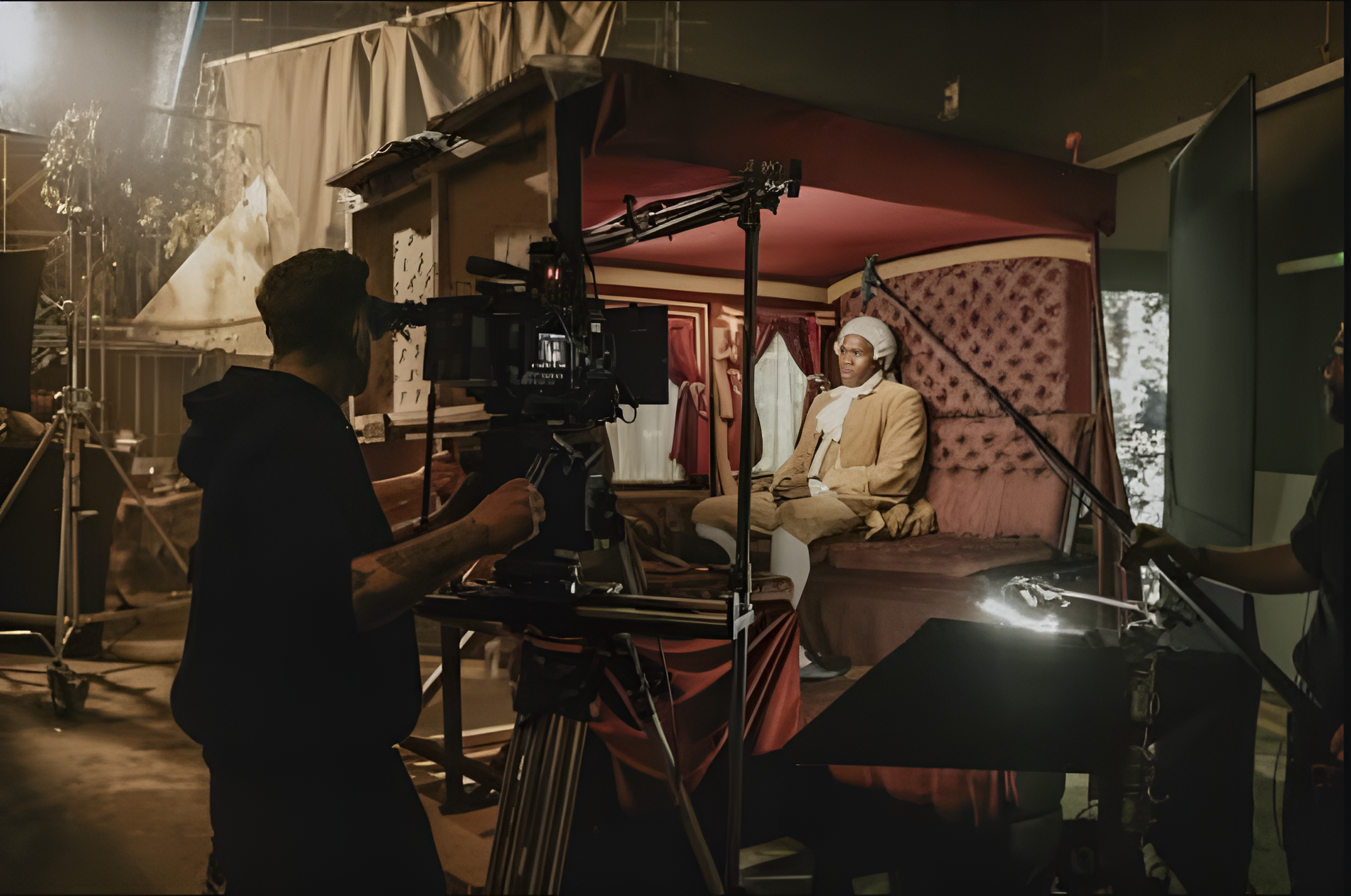 Behind the scenes of a film or photo shoot showing a man dressed in historical costume seated inside a vintage bed or carriage with a canopy, while crew members operate camera and equipment around him in a studio setting.