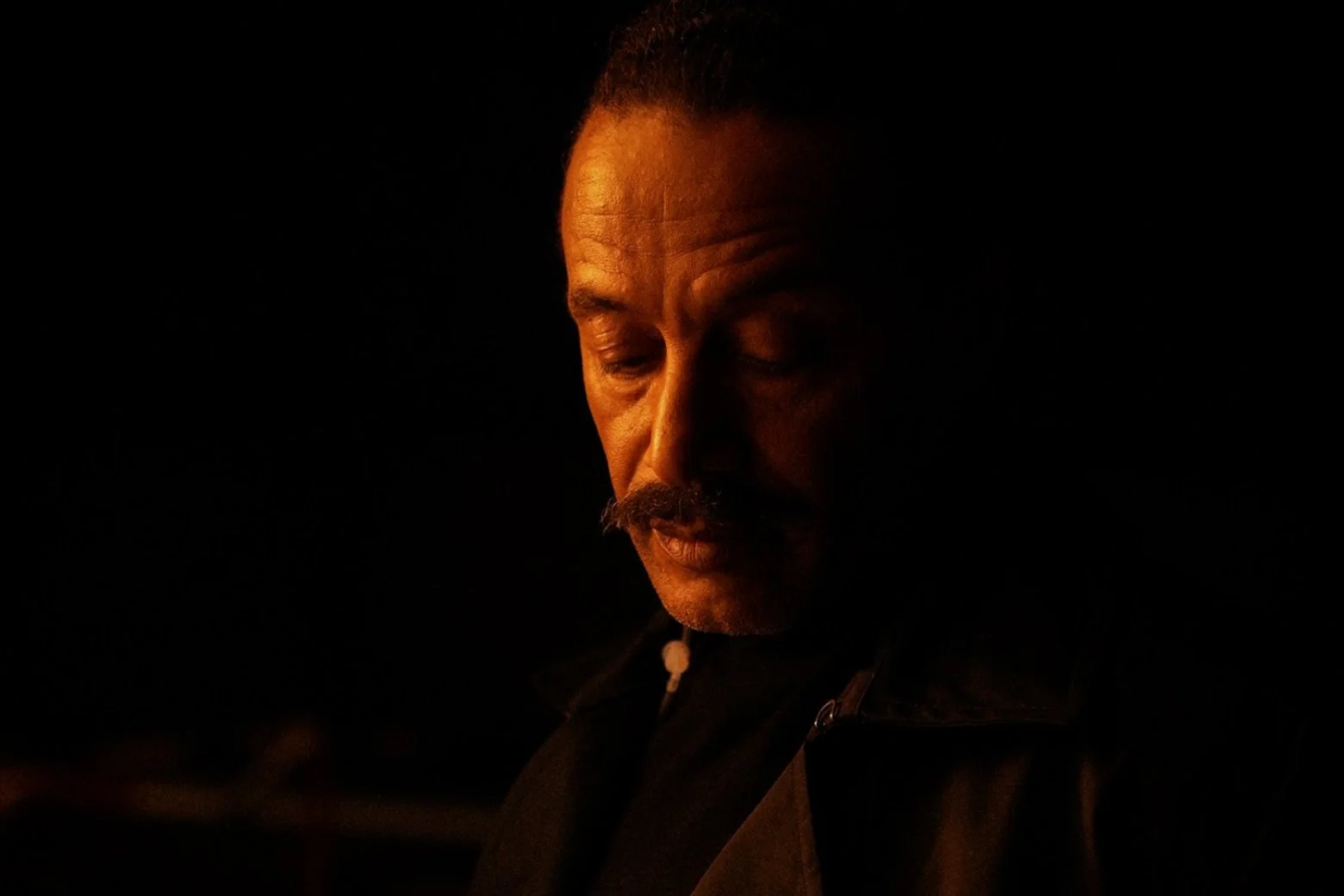 A man with a mustache and short hair looking downward in a dark setting with minimal lighting.