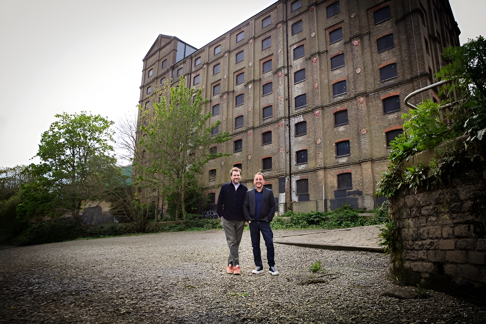 Two men standing outdoors in front of an old multi-story building with a brick exterior and many boarded-up windows. The men are smiling, dressed in semi-formal attire, and standing on a gravel surface near some greenery and trees.