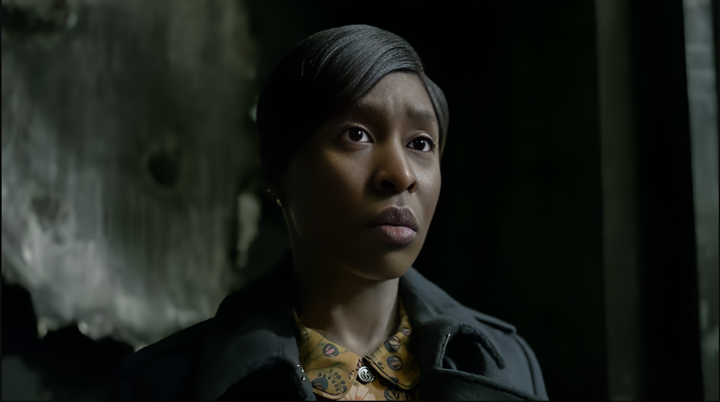 A young woman with dark skin and short dark hair looking directly at the camera in a dimly lit environment.