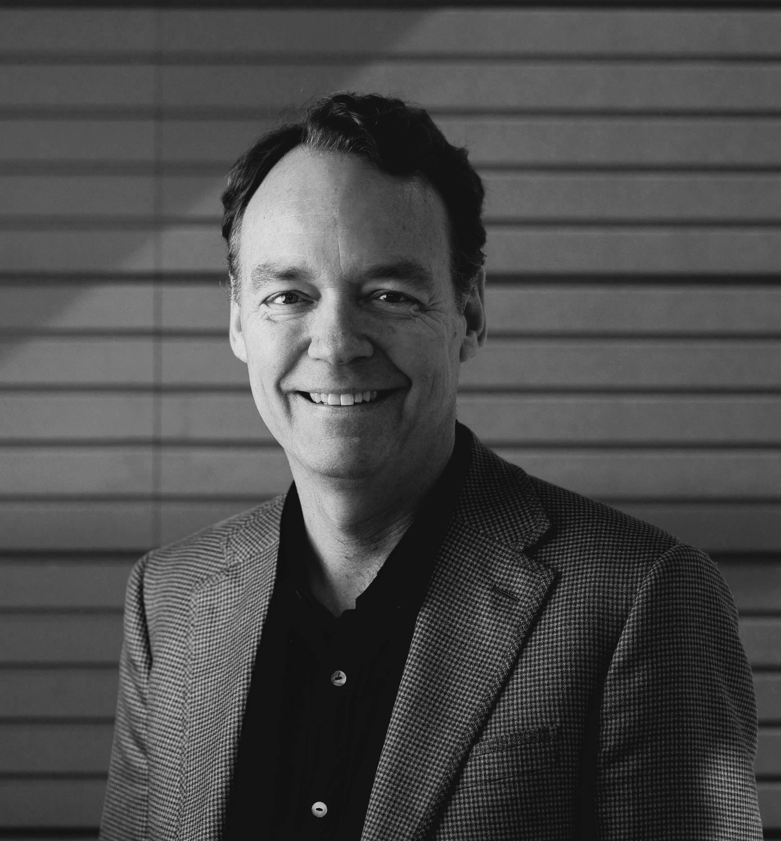 Black and white photograph of a smiling man with dark hair and a checkered blazer, standing in front of a horizontally paneled wall.