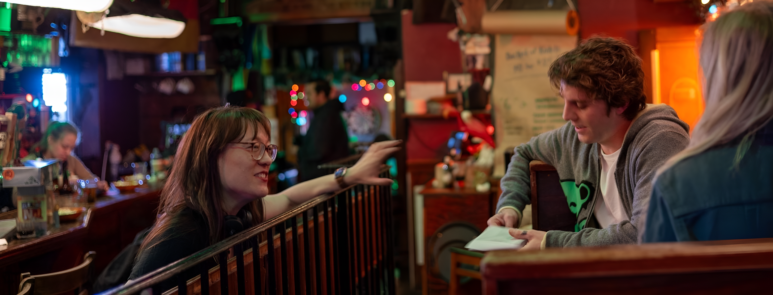 Three people sitting at a bar, engaging in conversation, with colorful lights and decorations in the background.