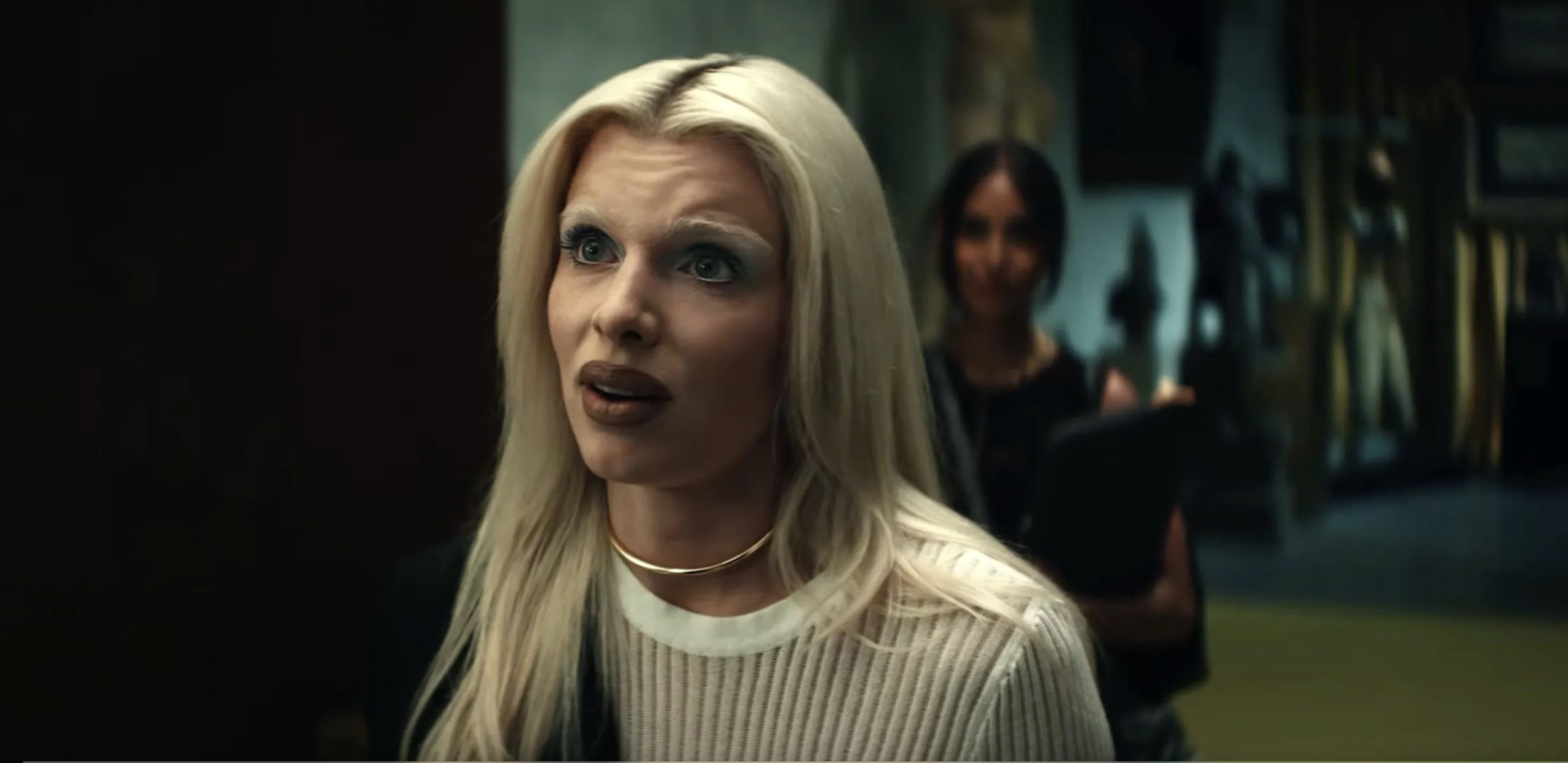 A woman with long blonde hair, wearing a white sweater and gold necklaces, appears surprised or concerned, looking to her left. In the background, there are blurred figures of women and a room with shelves and hanging objects.