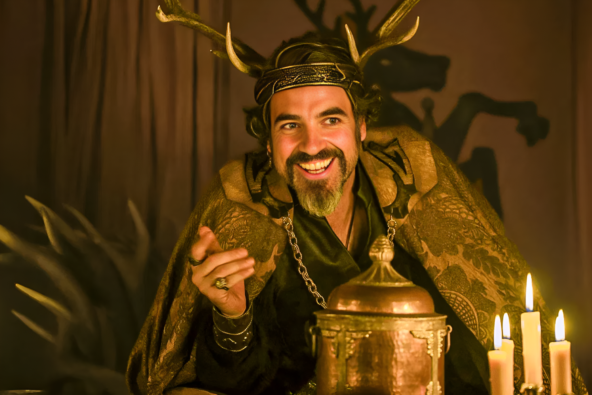 A man with a beard and long hair, wearing a crown with antlers, dressed in medieval or fantasy-style clothing, smiling, sitting at a table with lit candles in a dimly lit setting.