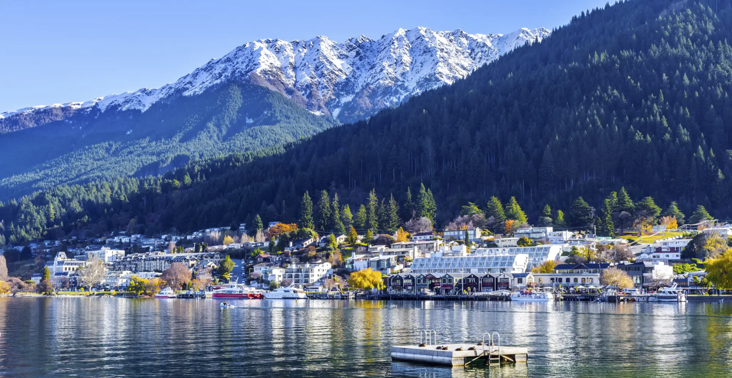 A lakeside town at the foot of snow-capped mountains, with boats on the water and trees with fall foliage.