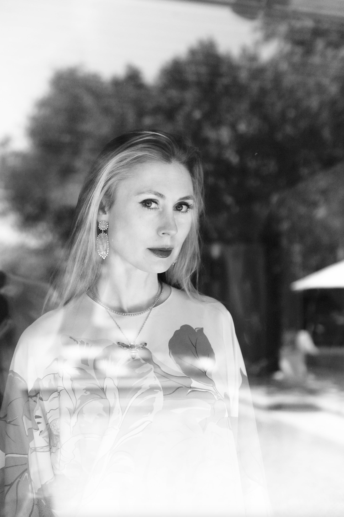 A black-and-white photo of a woman with long hair, wearing earrings and a necklace, in an outdoor setting with trees in the background.