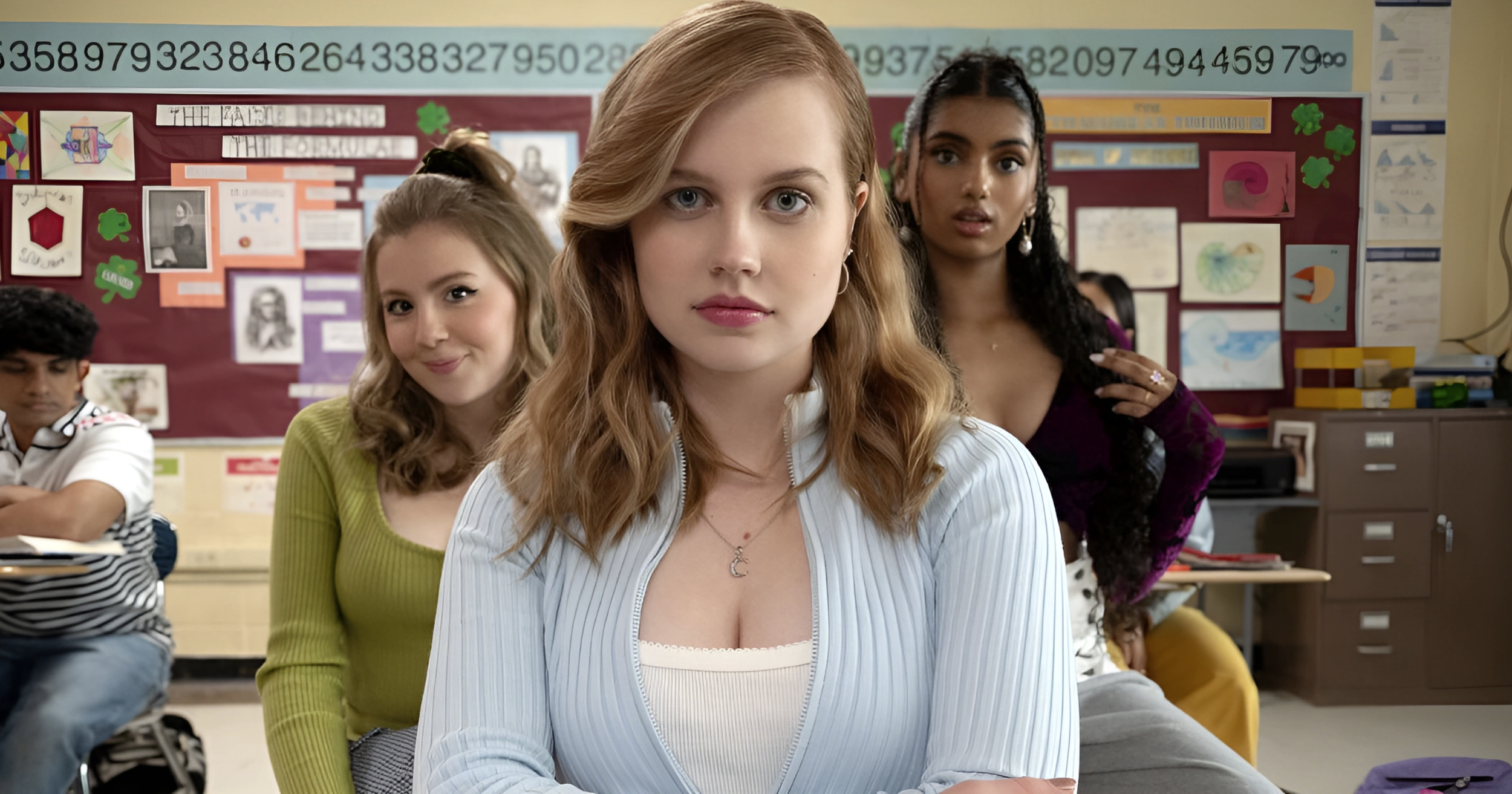 A group of students, primarily young women, in a classroom, with a girl with long, wavy red hair and a striped shirt in the center, looking directly at the camera, surrounded by classmates and educational decor on the walls.