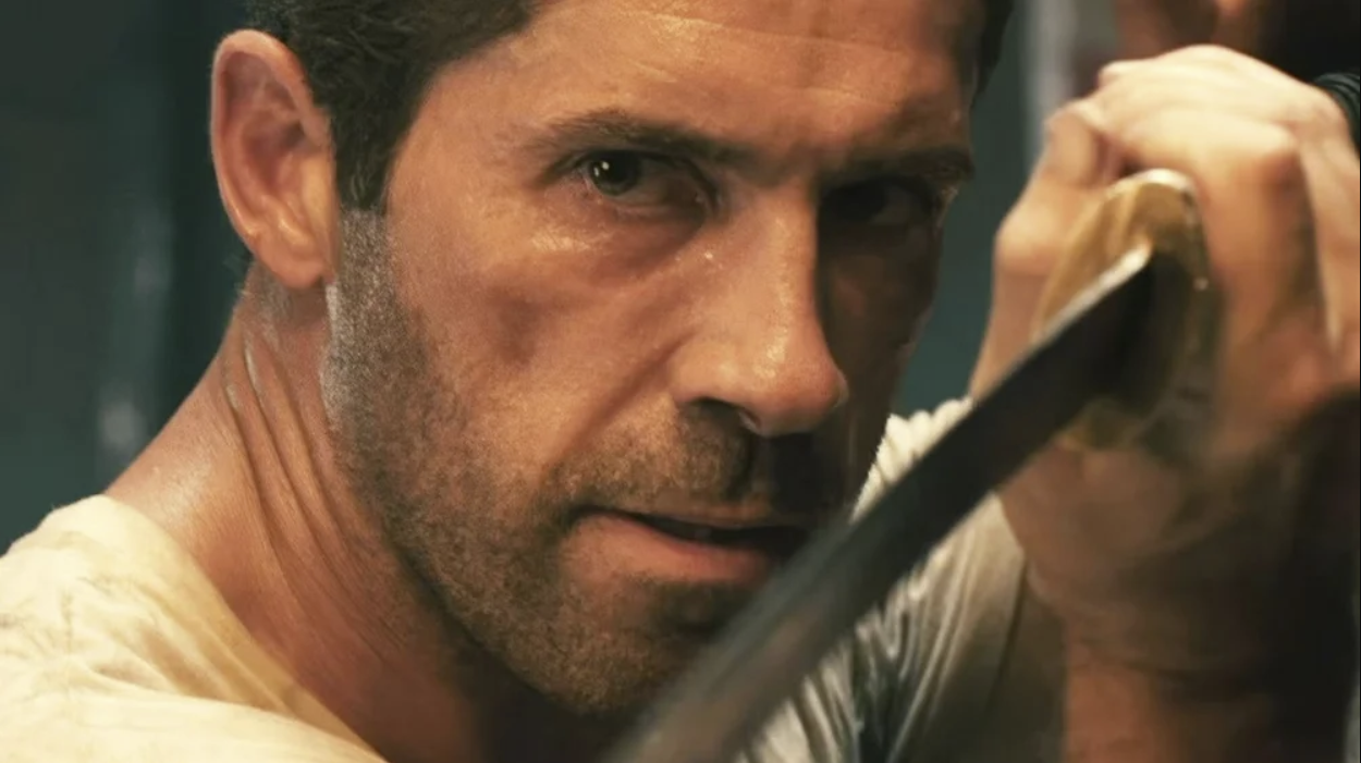 Close-up of a man with stubble, holding a sword with a determined expression, intense gaze, in a dimly lit setting.