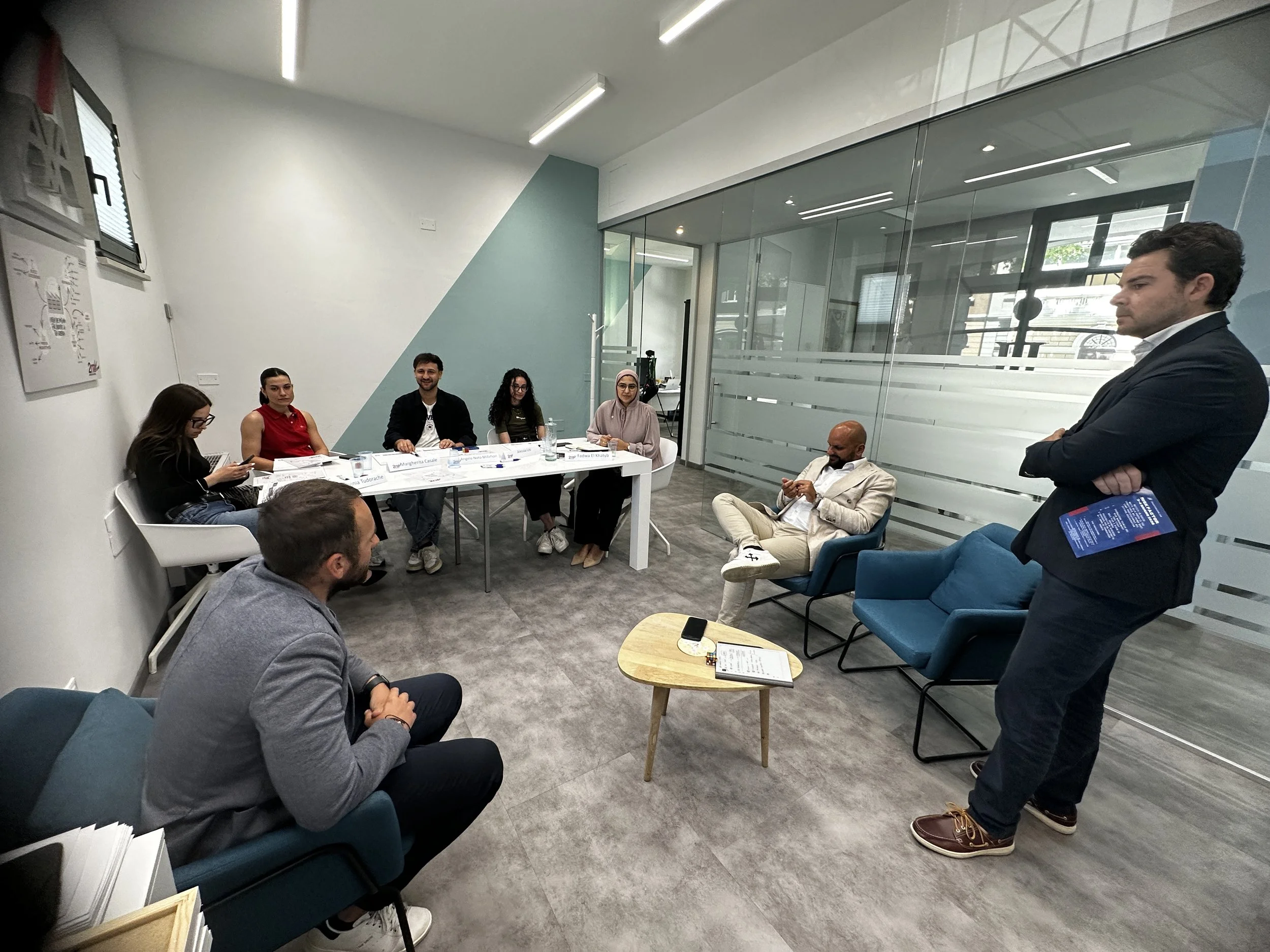 Un gruppo di persone si riunisce in una sala moderna con pareti bianche e vetrate, inquadrando alcuni dei partecipanti che sono seduti e uno che sta alzando le braccia.
