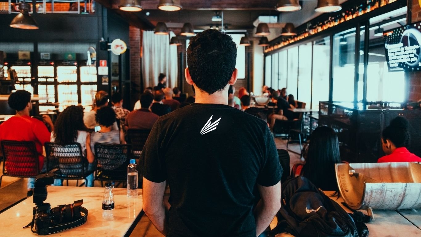 Immagine di un bar o ristorante con molte persone sedute ai tavoli, vista dalla parte di una persona con capelli corti, che indossa una maglietta nera, che si trova di fronte a un pubblico impegnato in una presentazione o evento.