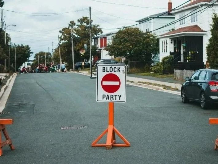 Segnale di blocco con scritto 'BLOCK PARTY' sotto il simbolo di divieto di accesso, bloccando la strada in una zona residenziale.