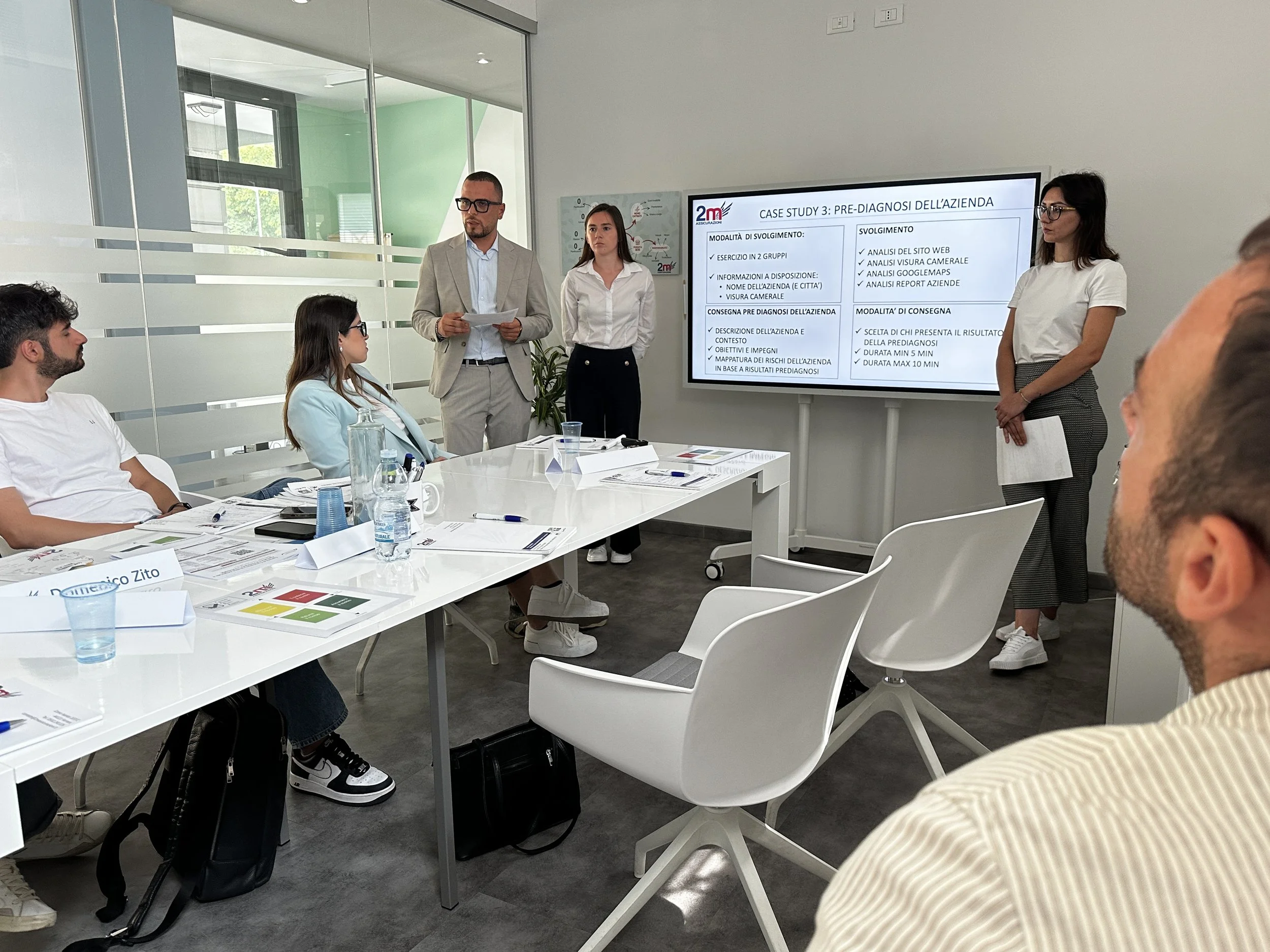 Presentazione in una sala di riunioni con cinque persone. Sullo schermo grande si vede un diagramma con testo in italiano, alcuni elemnti di una presentazione su una strategia aziendale o analisi di un caso. Il pubblico è seduto attorno a un tavolo con documenti e bottiglie d'acqua, mentre alcuni relatori stanno in piedi accanto allo schermo.