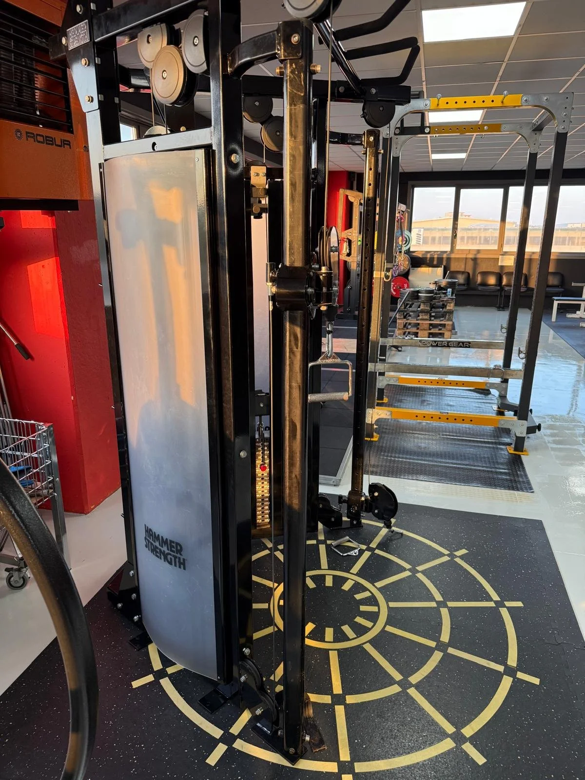 Attrezzo da palestra per allenamento di forza, con pilastri metallici, piastre di peso e una piattaforma di esercizio, in una palestra ben illuminata con grandi finestre.