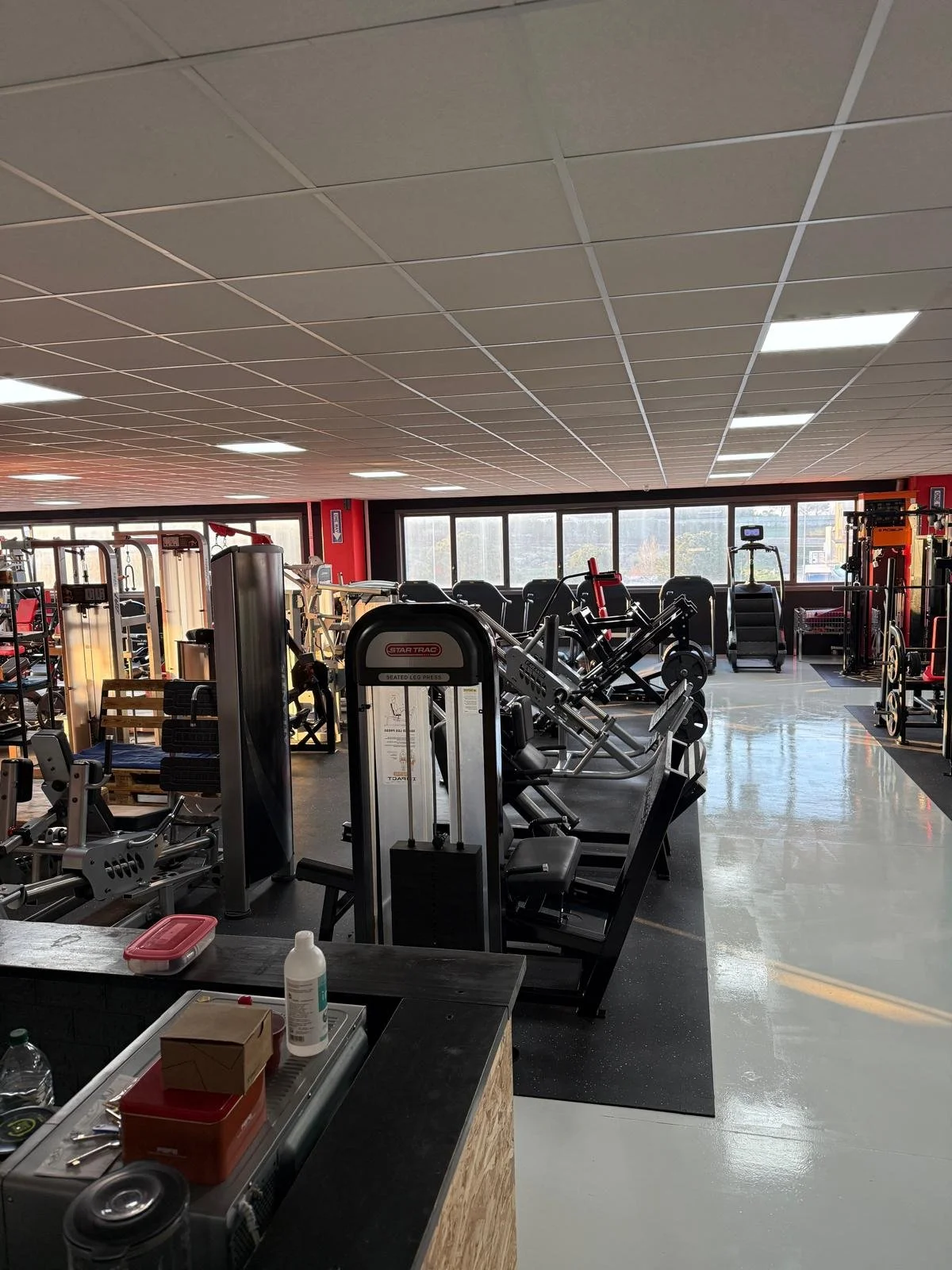 Interno di una palestra con attrezzature come pesi, tapis roulant e macchinari da allenamento, illuminata dalla luce naturale attraverso grandi finestre.