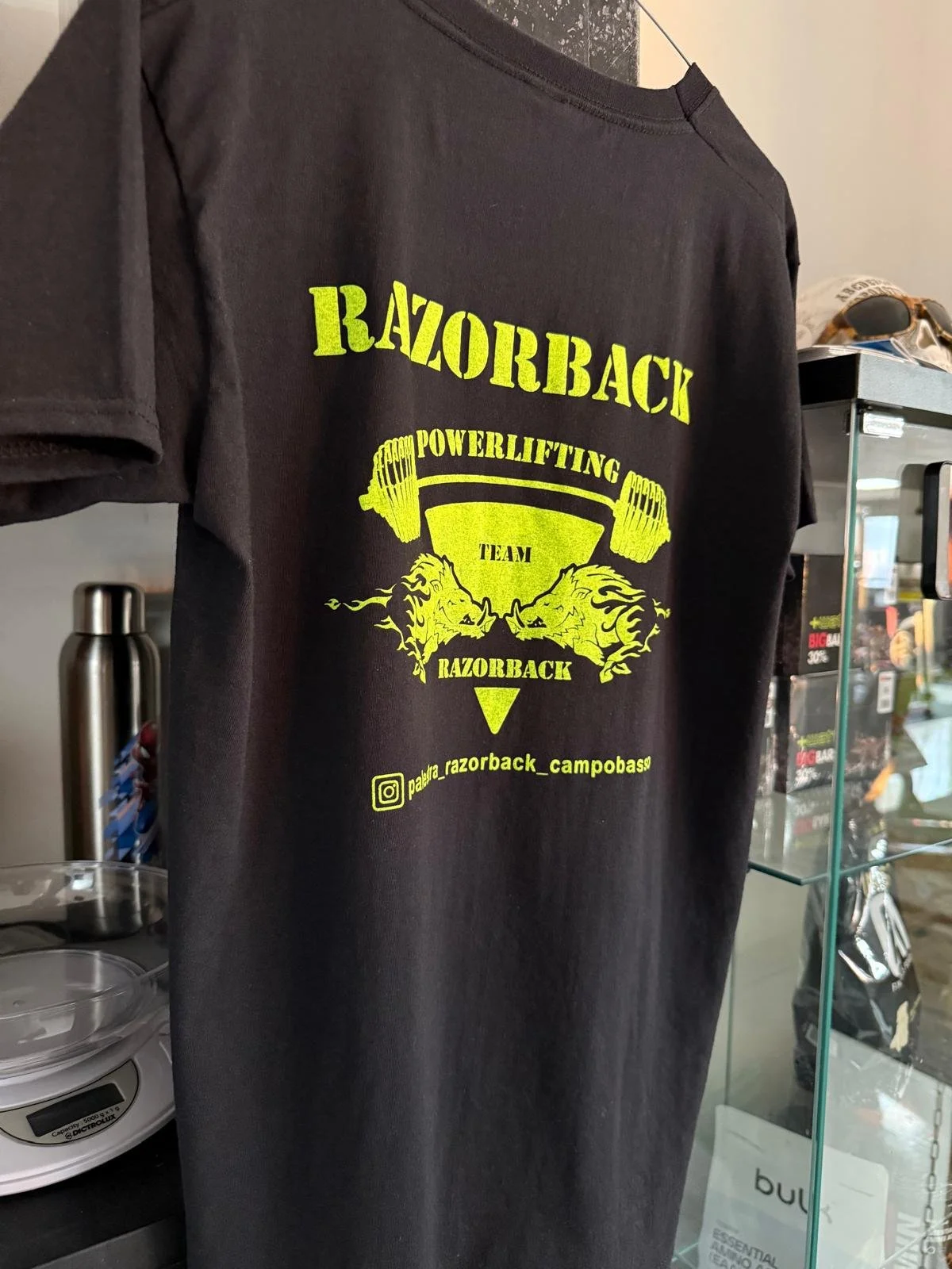 Maglietta nera con logo giallo di Razorback Powerlifting Team, dove si vedono un bilanciere con due leoni stilizzati e il testo 'RAZORBACK POWERLIFTING TEAM' e il loro profilo Instagram.