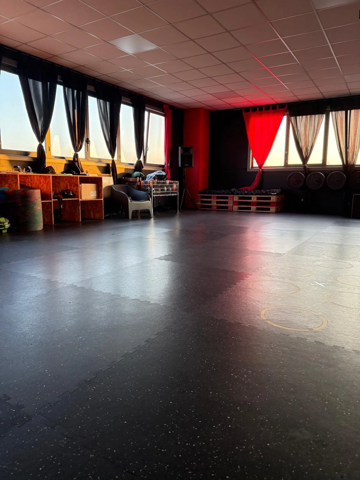 Sala di danza con pavimento in gomma, pallet di attrezzi e sedie, con grandi finestre e tende nere e rosse.