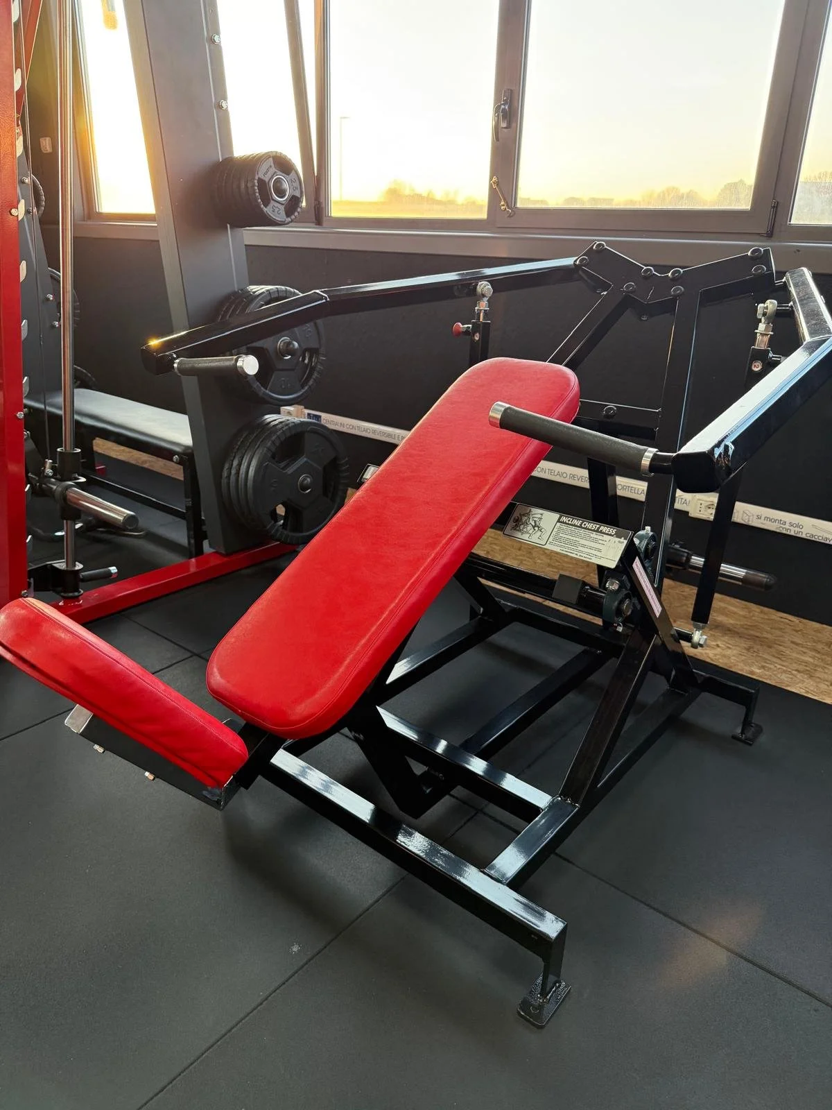 Attrezzatura da palestra con panca regolabile con schienale rosso e altra macchina per esercizi, vicino a una finestra con vista verso l'esterno.