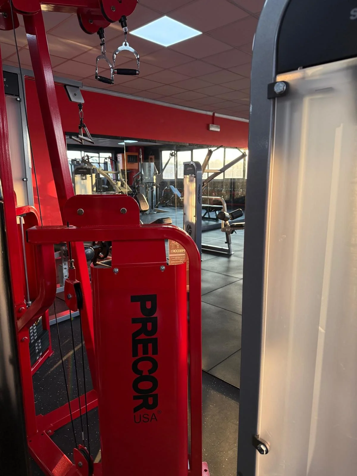 Attrezzature da palestra in un centro fitness, tra cui una macchina rossa con la scritta 'PRECOR USA' e altri attrezzi come manubri e macchinari, con grandi finestre che lasciano entrare la luce naturale.