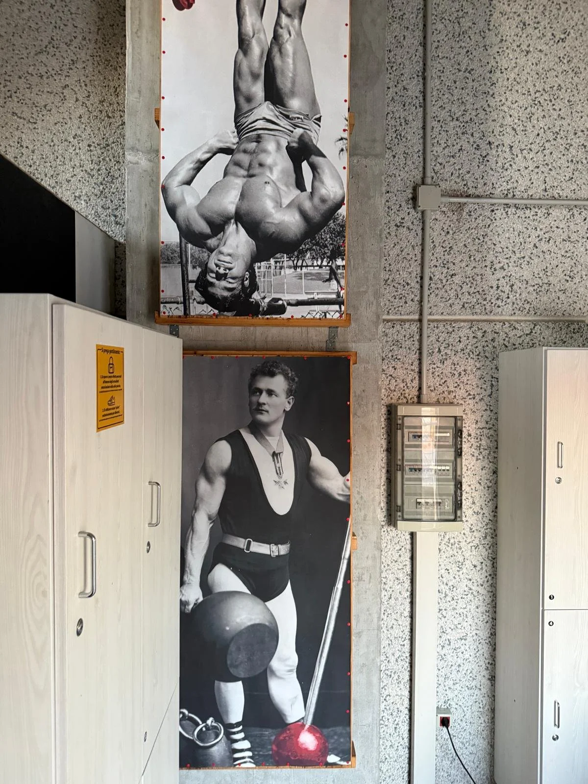 Due grandi poster in bianco e nero raffigurano uomini muscolosi: uno in posizione di allenamento con i muscoli in evidenza e l'altro con un manubrio e una palla da training, entrambi in ambienti interni.