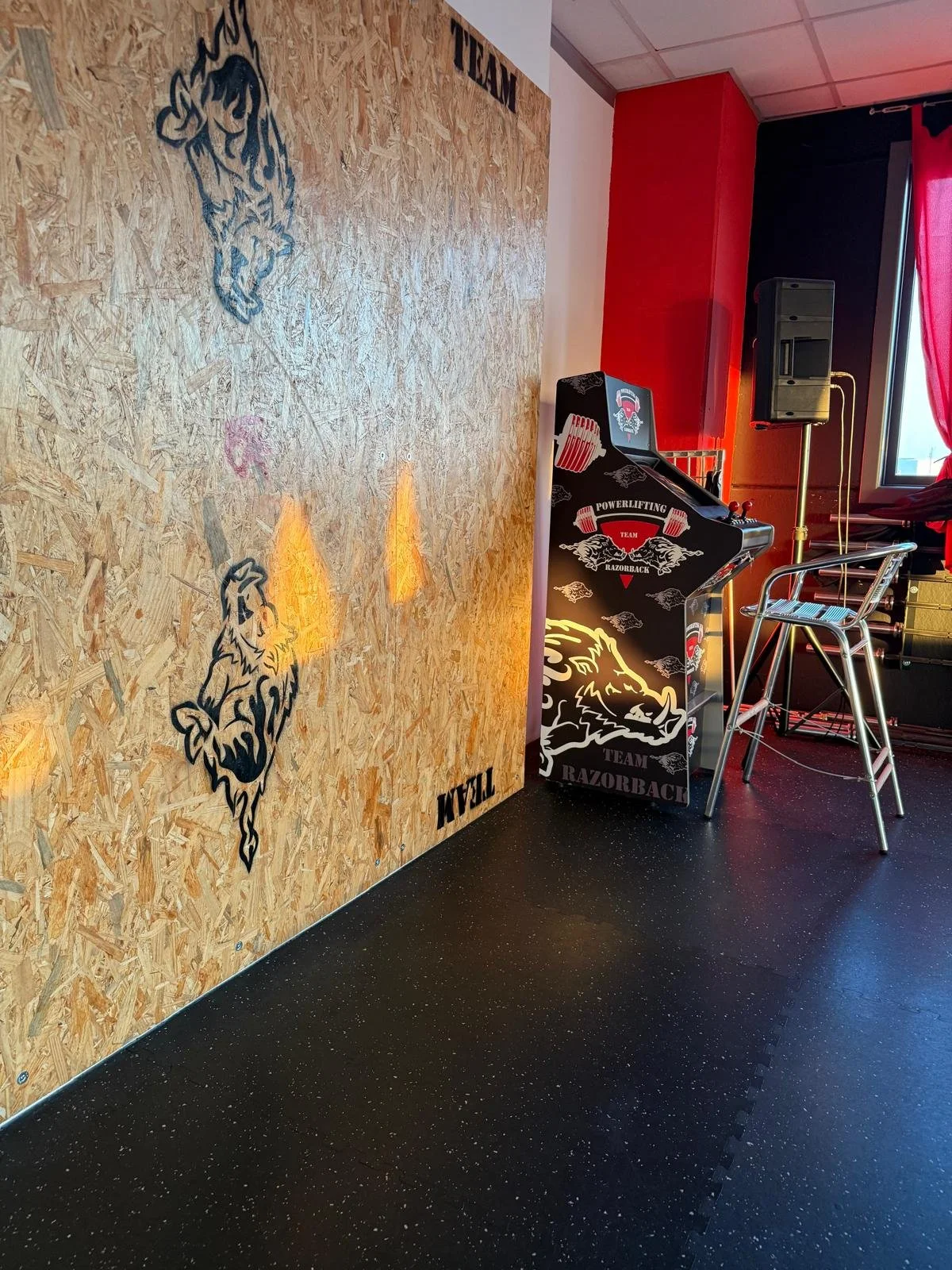 Parete di compensato con disegni di lupi e la scritta 'Well' e 'Titan'. Area con un piano di gioco da powerlifting, sedie di metallo, altoparlante e finestra con tende rosse.