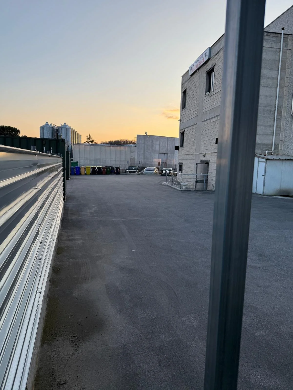 Un cortile industriale con parcheggio, si vedono vari veicoli e contenitori dei rifiuti, con un edificio moderno sullo sfondo al tramonto.