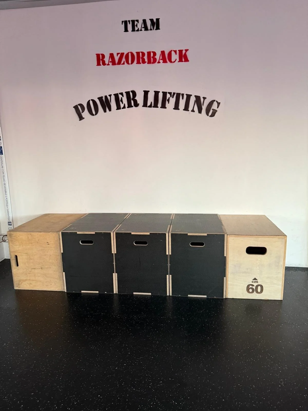 Set di scatole in legno e metallo usate come attrezzi in palestra, con l'etichetta di 60 cm di altezza, disposte sul pavimento di un centro fitness. Sul muro dietro, scritte in italiano su sfondo bianco: 'TEAM RAZORBACK POWER LIFTING'.
