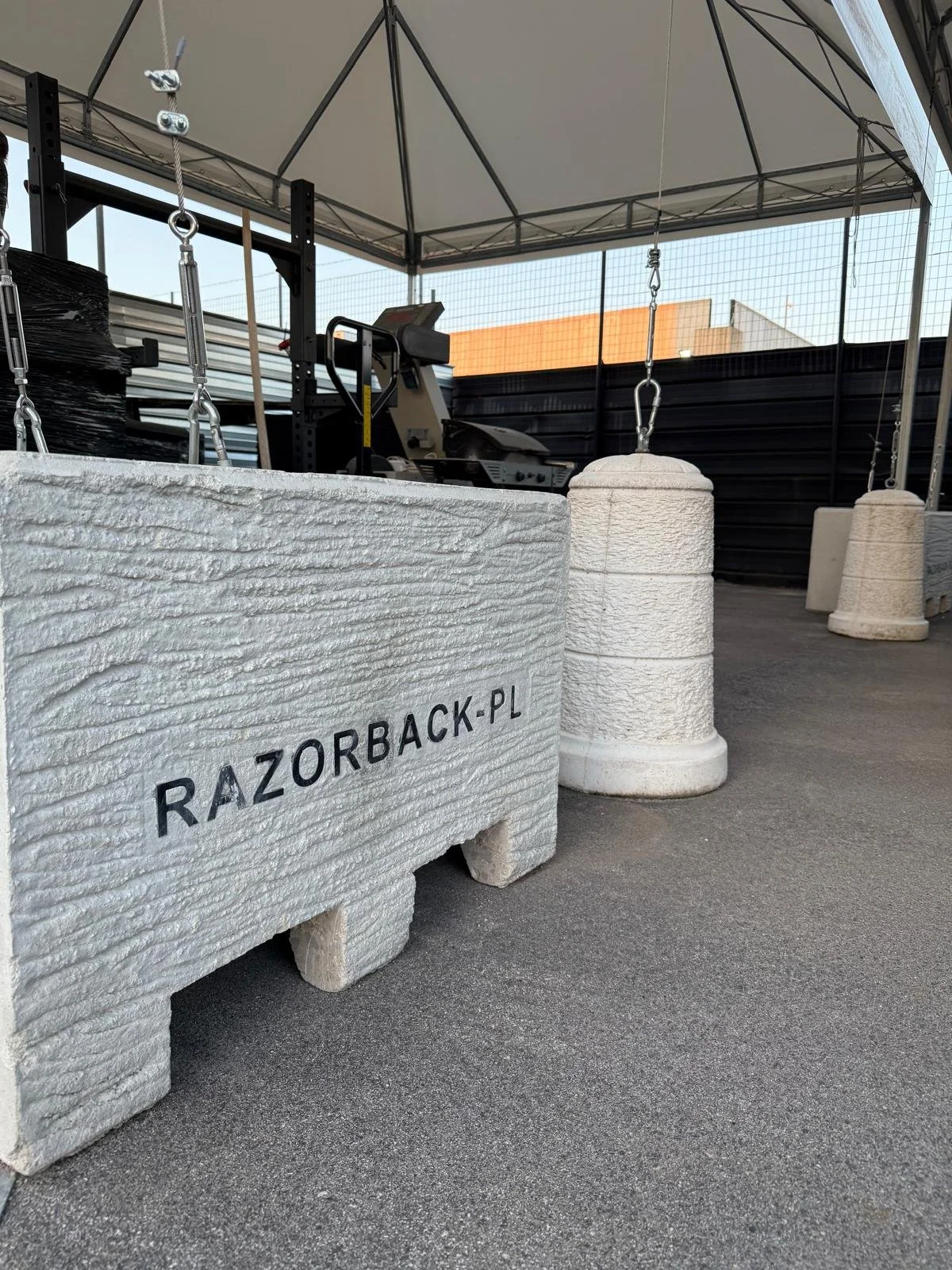 Foto di un'area all'aperto con panchine di cemento e attrezzature da palestra, tra cui un tapis roulant, sotto una struttura con tetto. Sul primo contenitore di cemento si legge "RAZORBACK.PL".