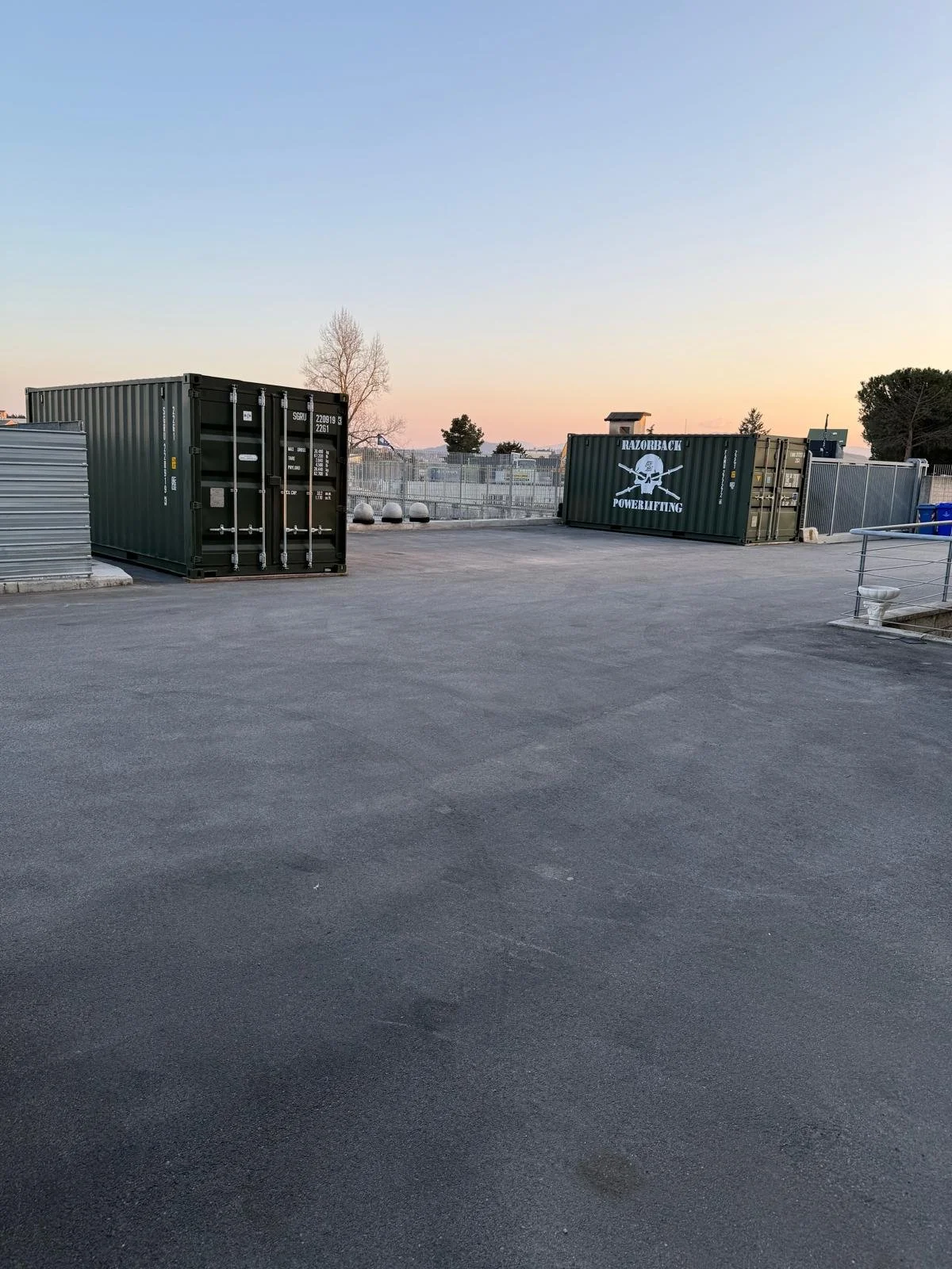 Sfondi di container da trasporto su un parcheggio all'aperto durante il tramonto, con una recinzione in fondo e alberi sullo sfondo.